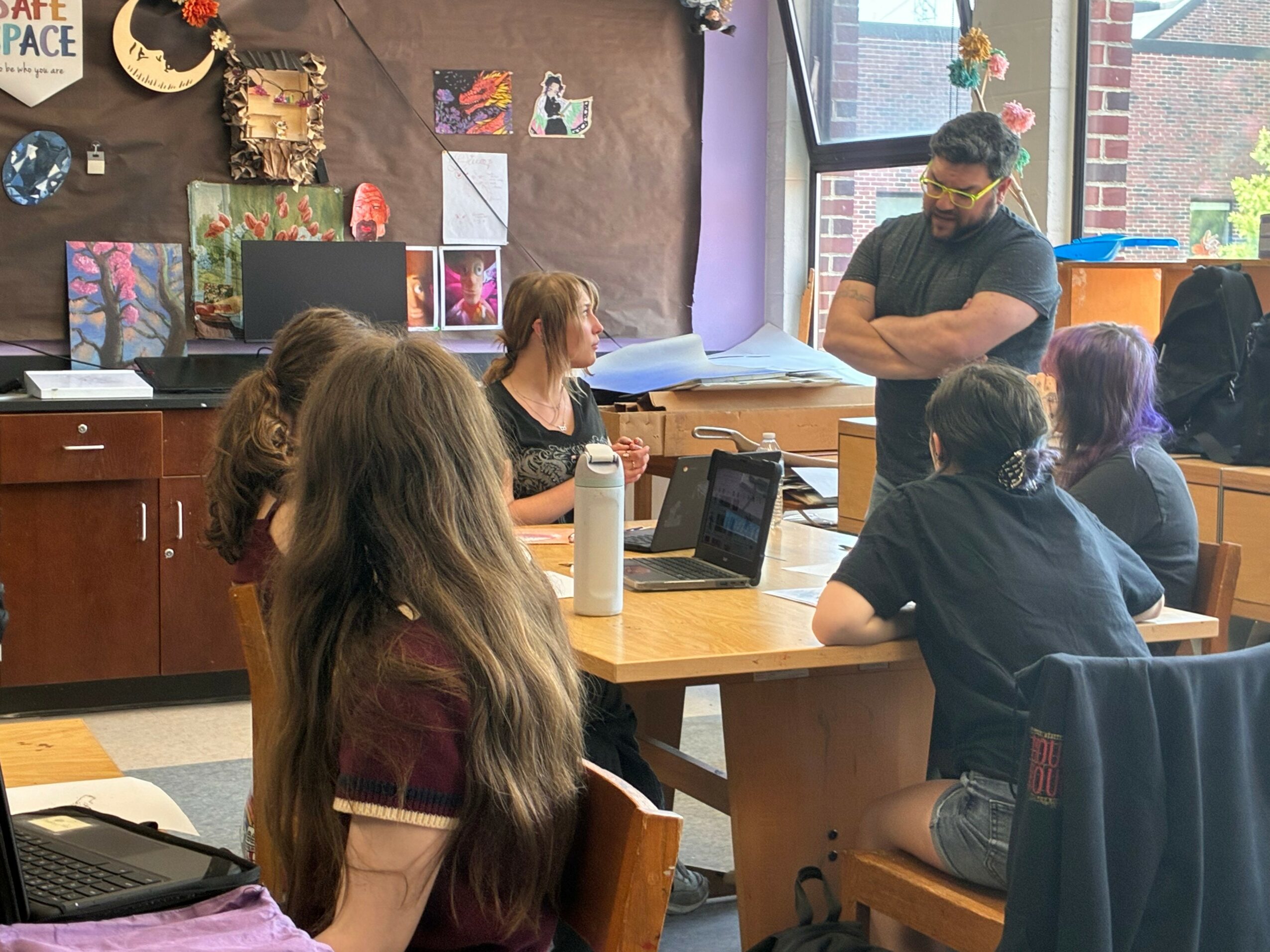 Teacher standing at a table with students in a classroom, colorful artwork on the wall, and a window in the background.