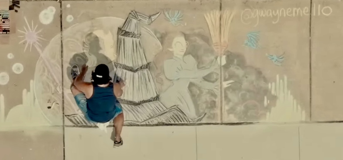 Child in blue shirt and hat drawing on sidewalk with chalk, colorful mural in background, overhead view.