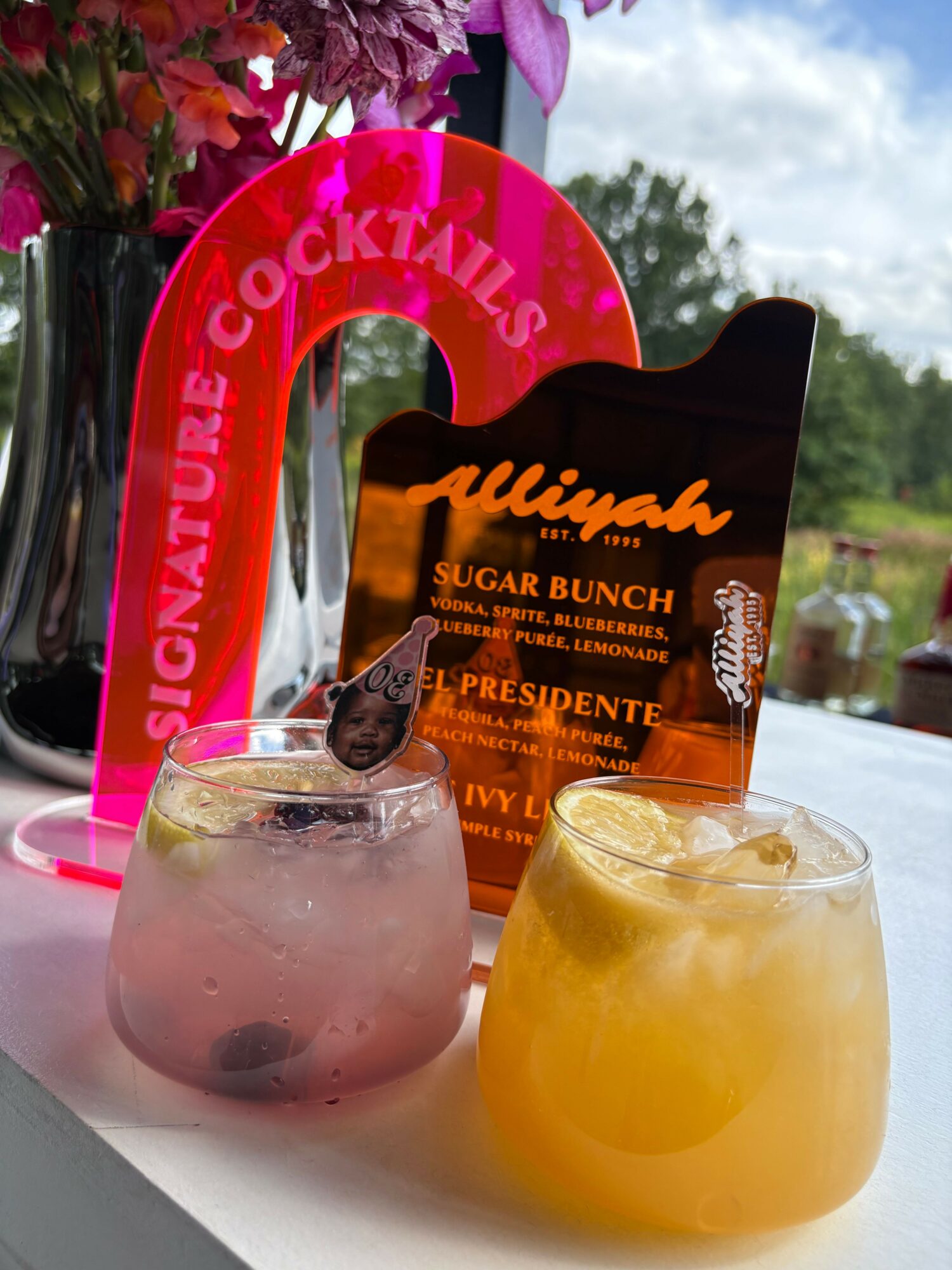 Two drinks on a table with a pink sign and flowers in the background, one yellow and one pink, with a black card.