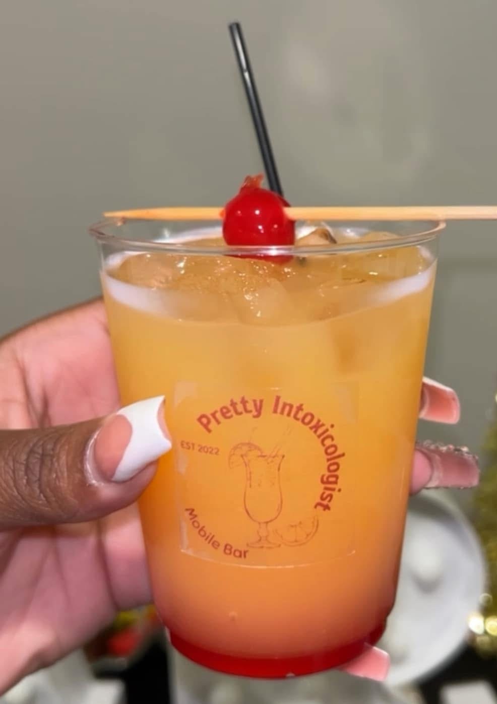 A colorful cocktail in a glass with a cherry and straw, held by a person with painted nails.