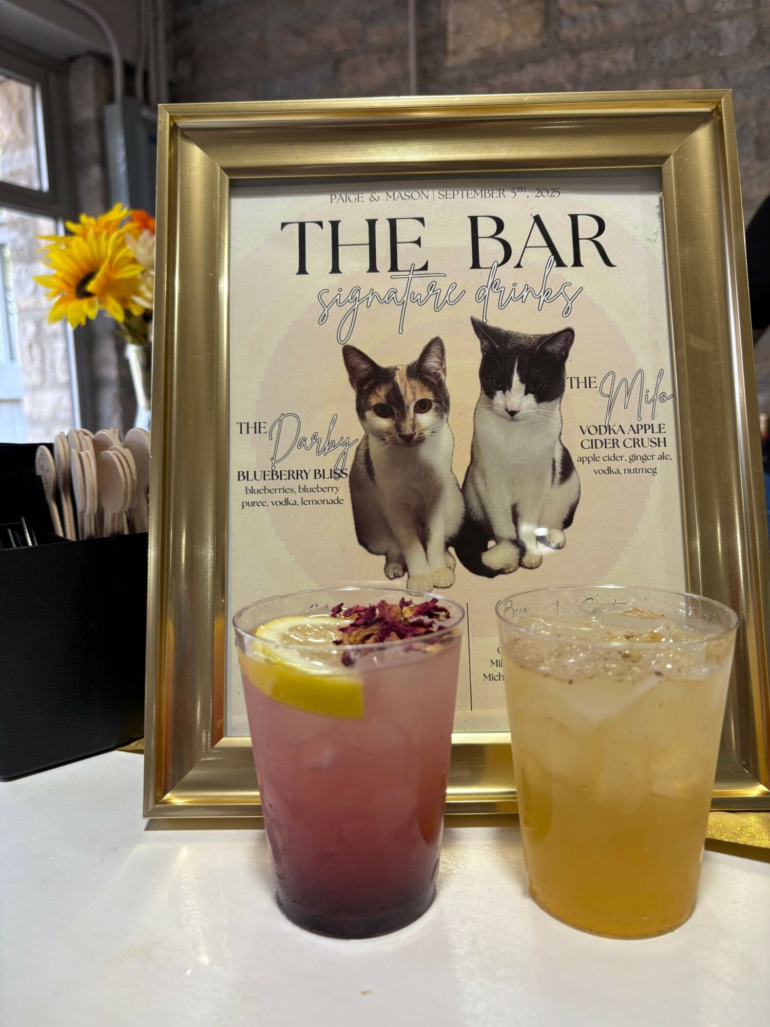 Framed poster advertising a dog-themed bar, with two dogs and text, placed on a table with two colorful drinks and a sunflower.
