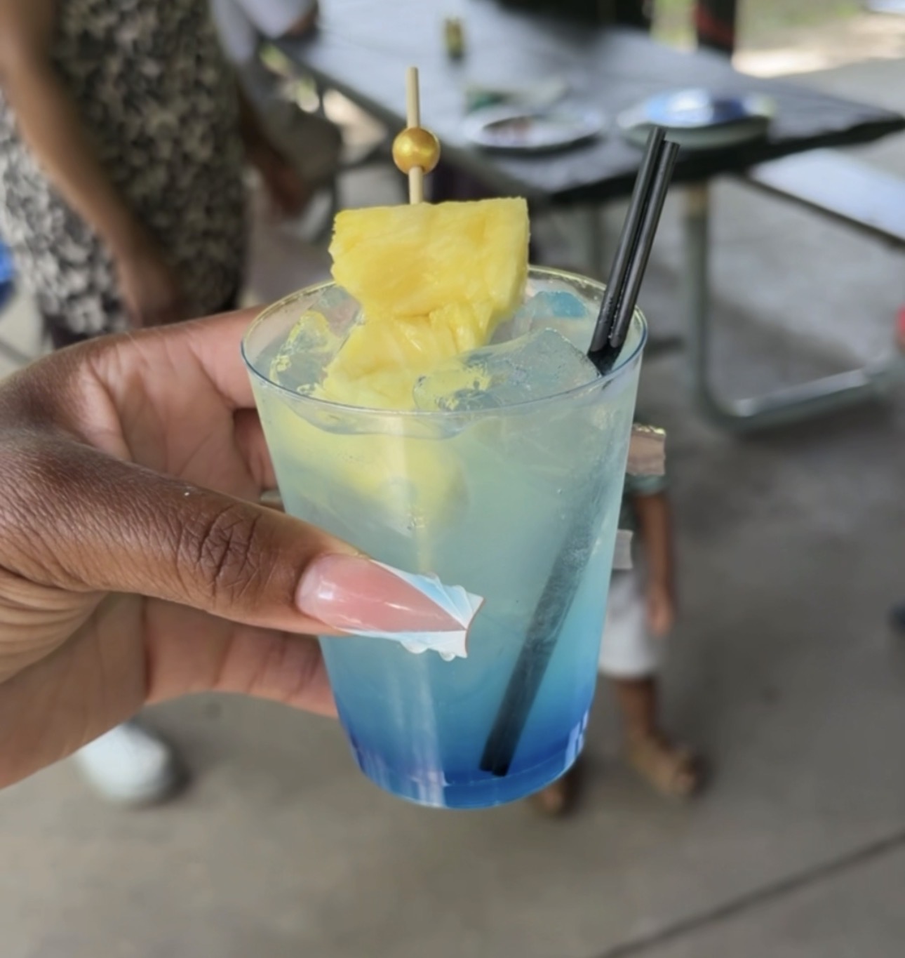 Hand holding a colorful layered drink with pineapple garnish and straw, outdoors setting, blurred background.