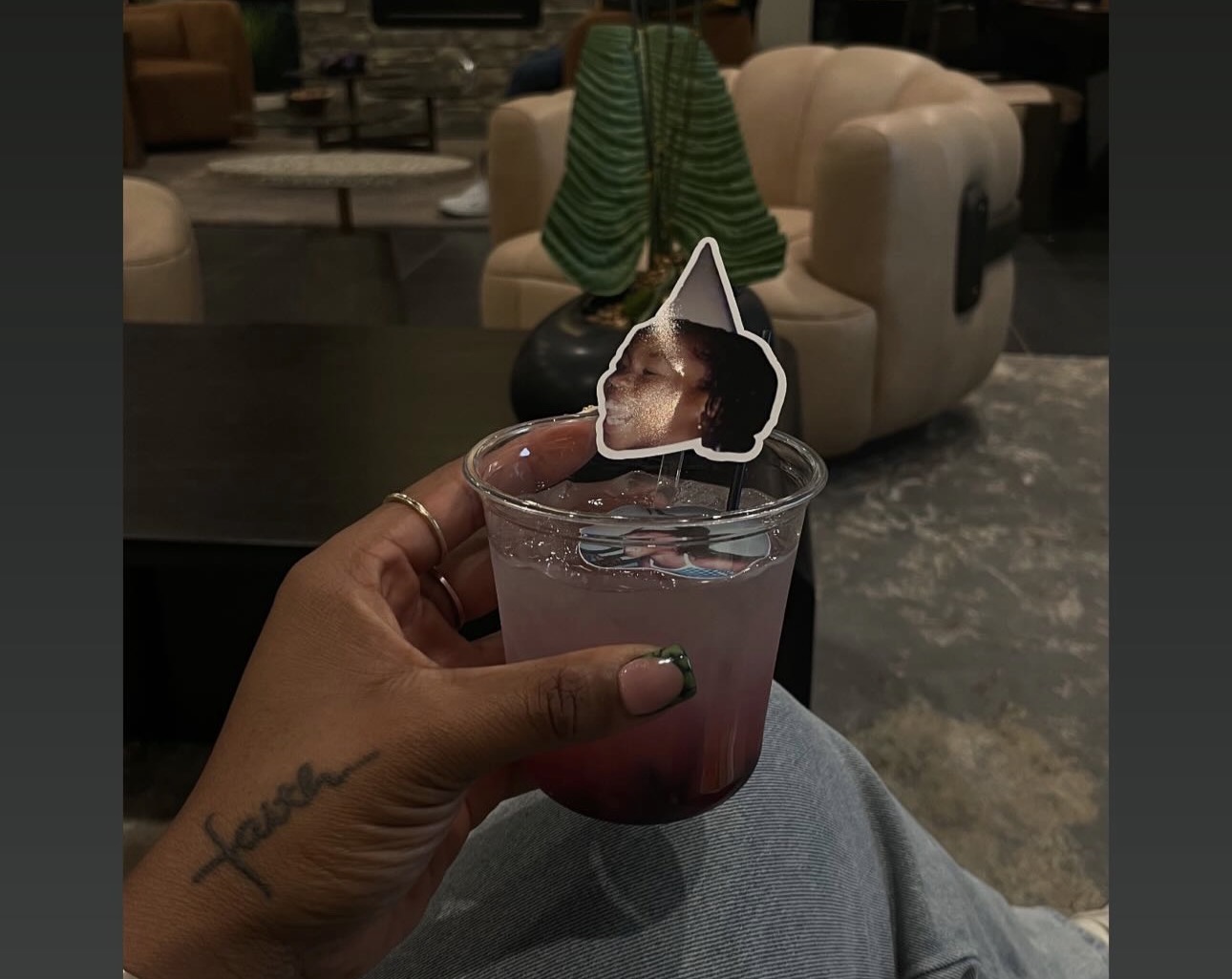 Person holding a drink with a small photo of a dog attached on top, in a cozy indoor setting.
