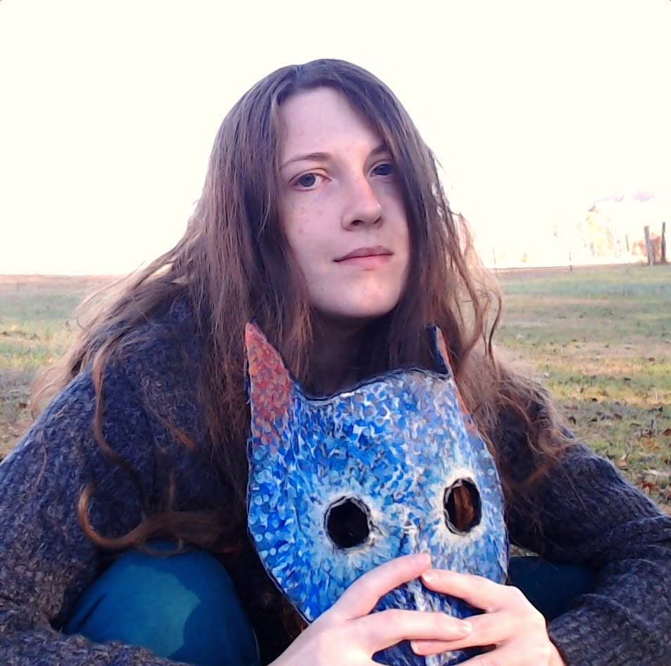 Young woman with long hair holding a blue owl-shaped mask outdoors, sitting on grass, with a field and sky in background.