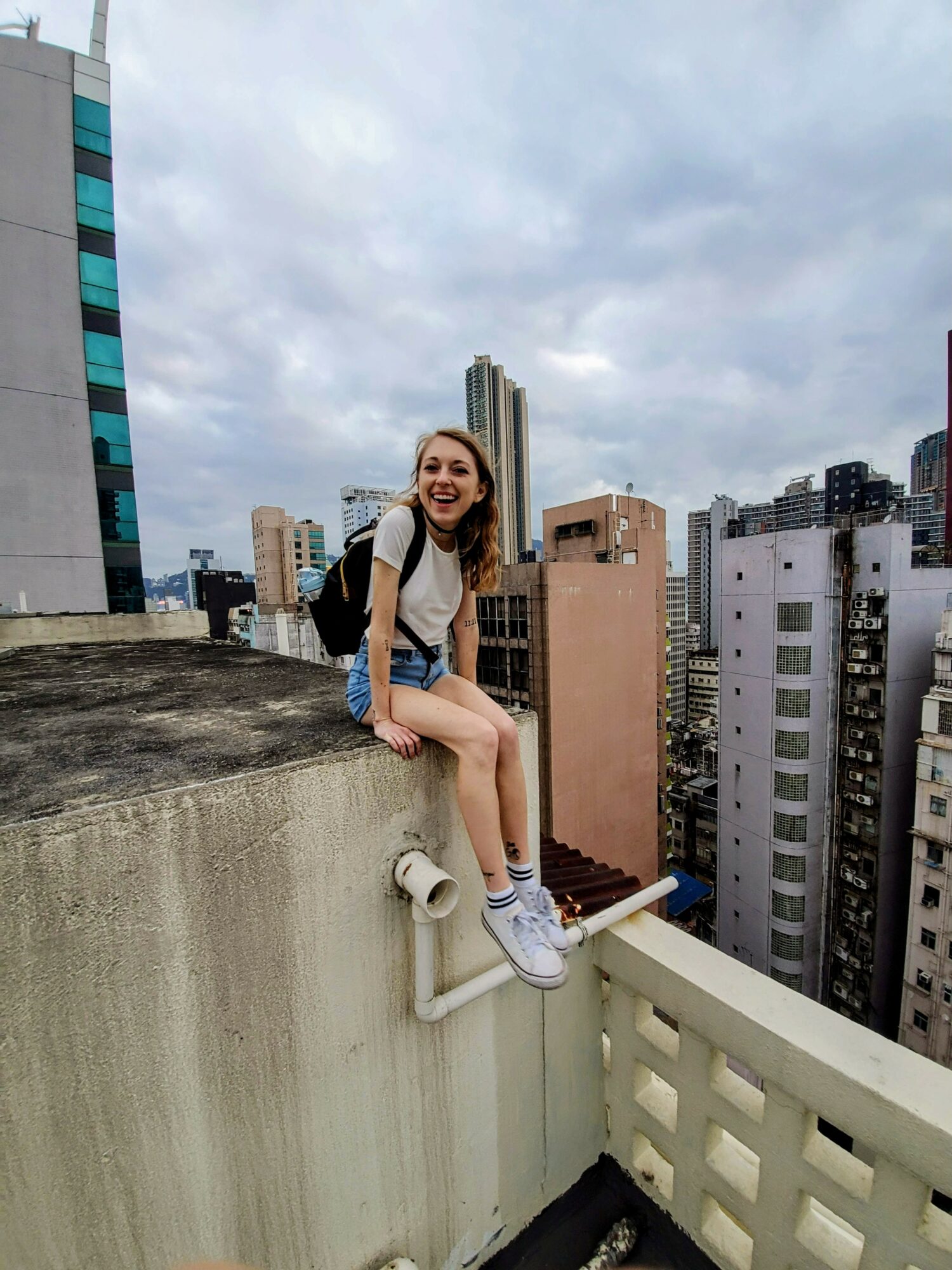 Young woman sitting on rooftop edge with city skyline behind her, smiling, wearing casual clothes and sneakers.