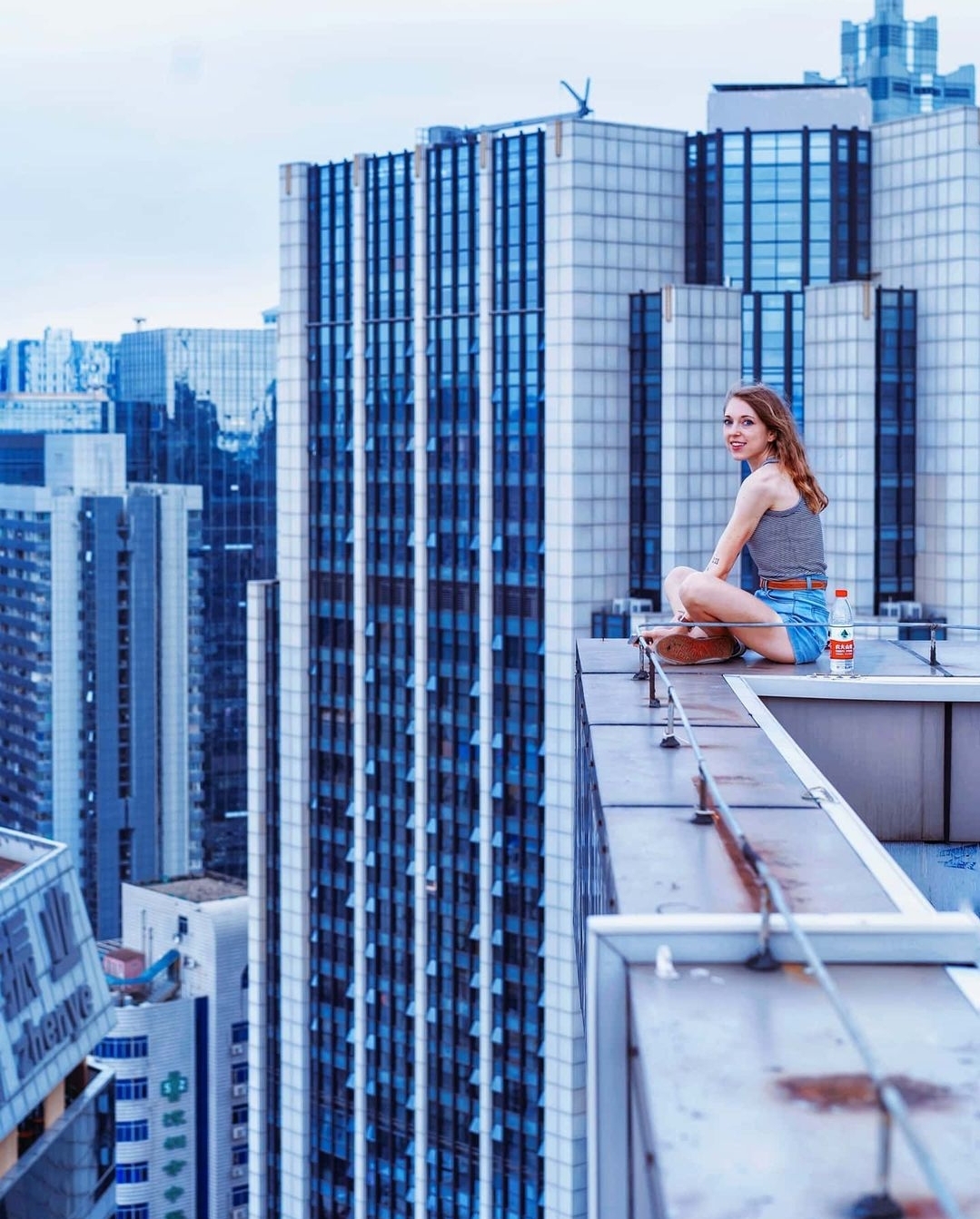 Woman sitting cross-legged on rooftop with city skyscrapers in background, blue sky, casual clothing, relaxed pose.