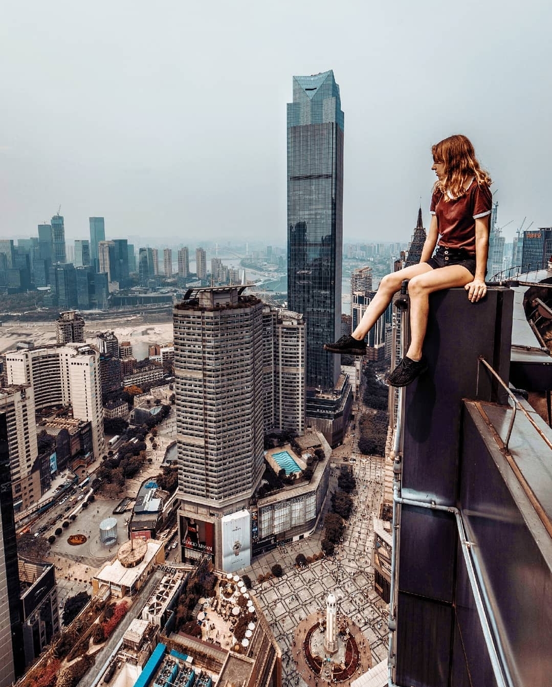 Woman sitting on rooftop edge overlooking city skyline with tall buildings and skyscrapers, cloudy sky, urban scene, daytime.