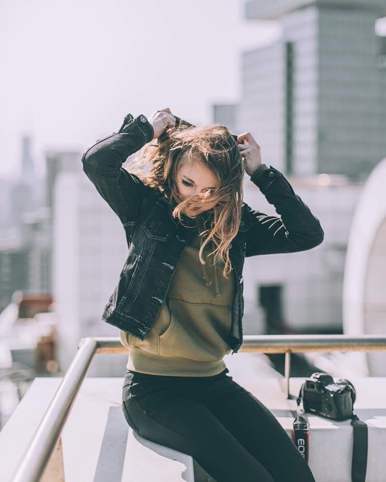 Young woman with long hair adjusting her hair outdoors, city buildings in background, wearing a black jacket and green top.
