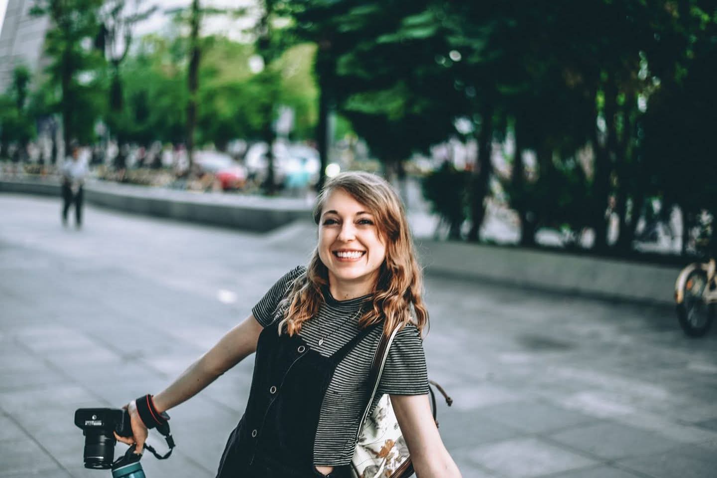 Smiling woman with shoulder-length hair, wearing a dark shirt and gray cardigan, standing outdoors on a city street.