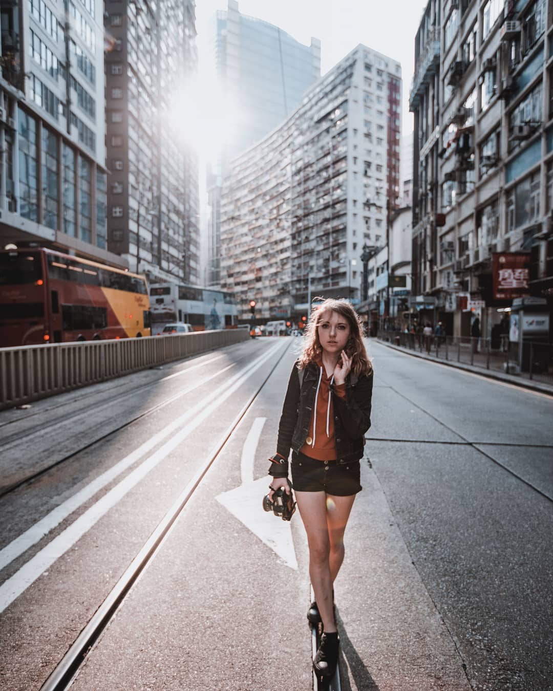 Young woman walking on city street with tall buildings, sunlight, and tram tracks, holding a camera, wearing a jacket and shorts.