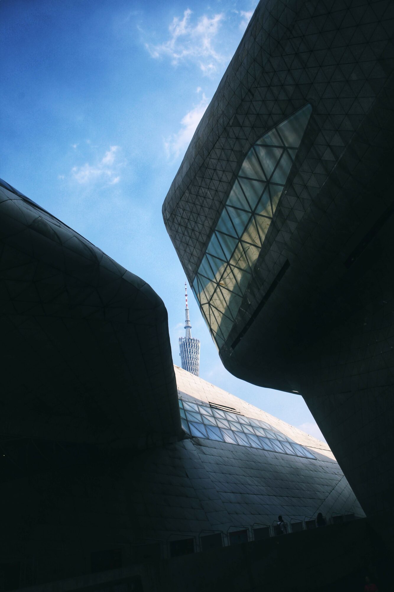 View of a tall building with a glass facade, sky with clouds, and a tower in the background, seen from below.