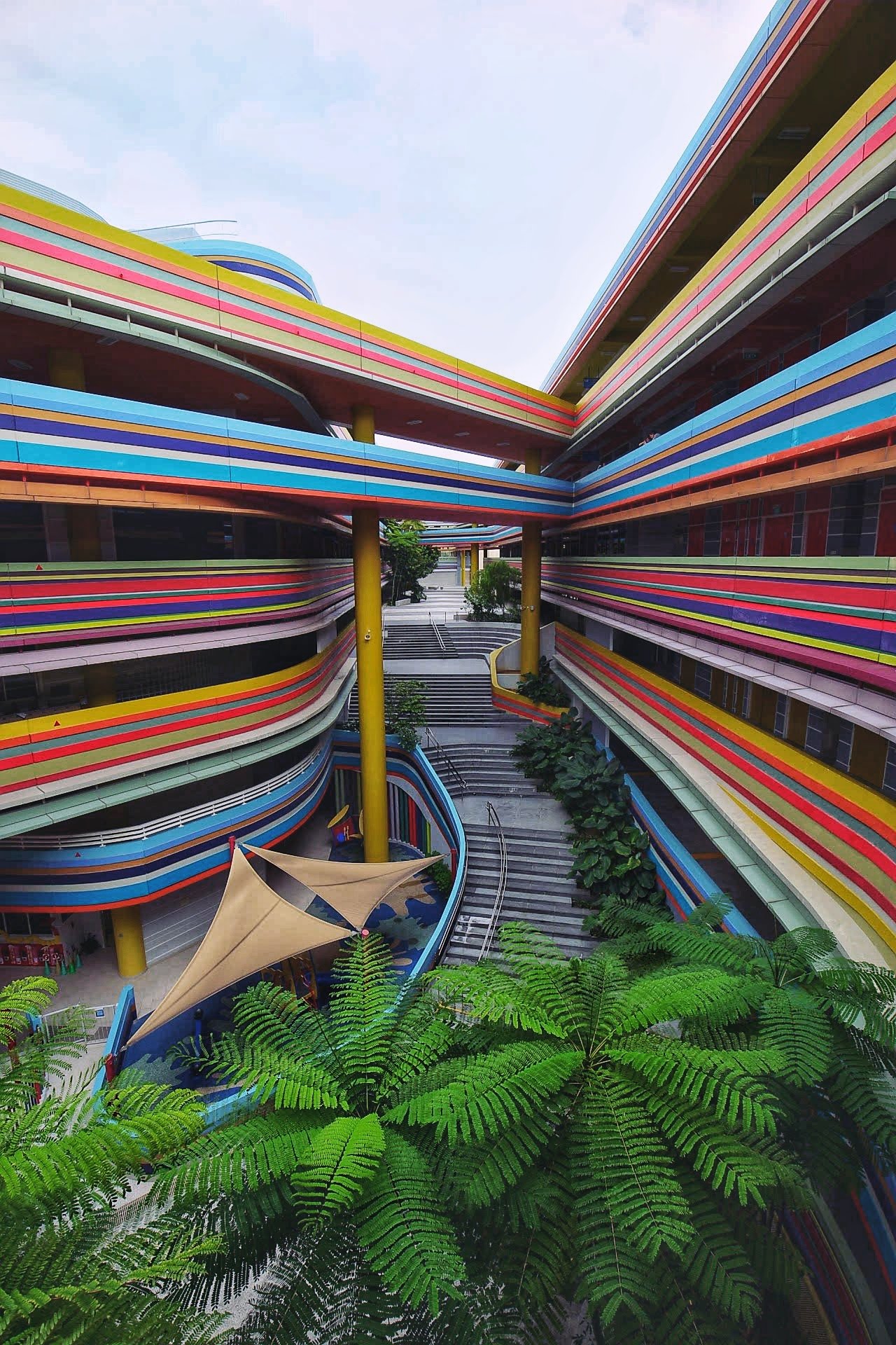 Colorful modern building with curved lines, greenery, and a walkway, viewed from below with a clear sky.