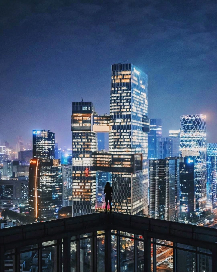 Person standing on rooftop overlooking a city skyline at night with illuminated skyscrapers.