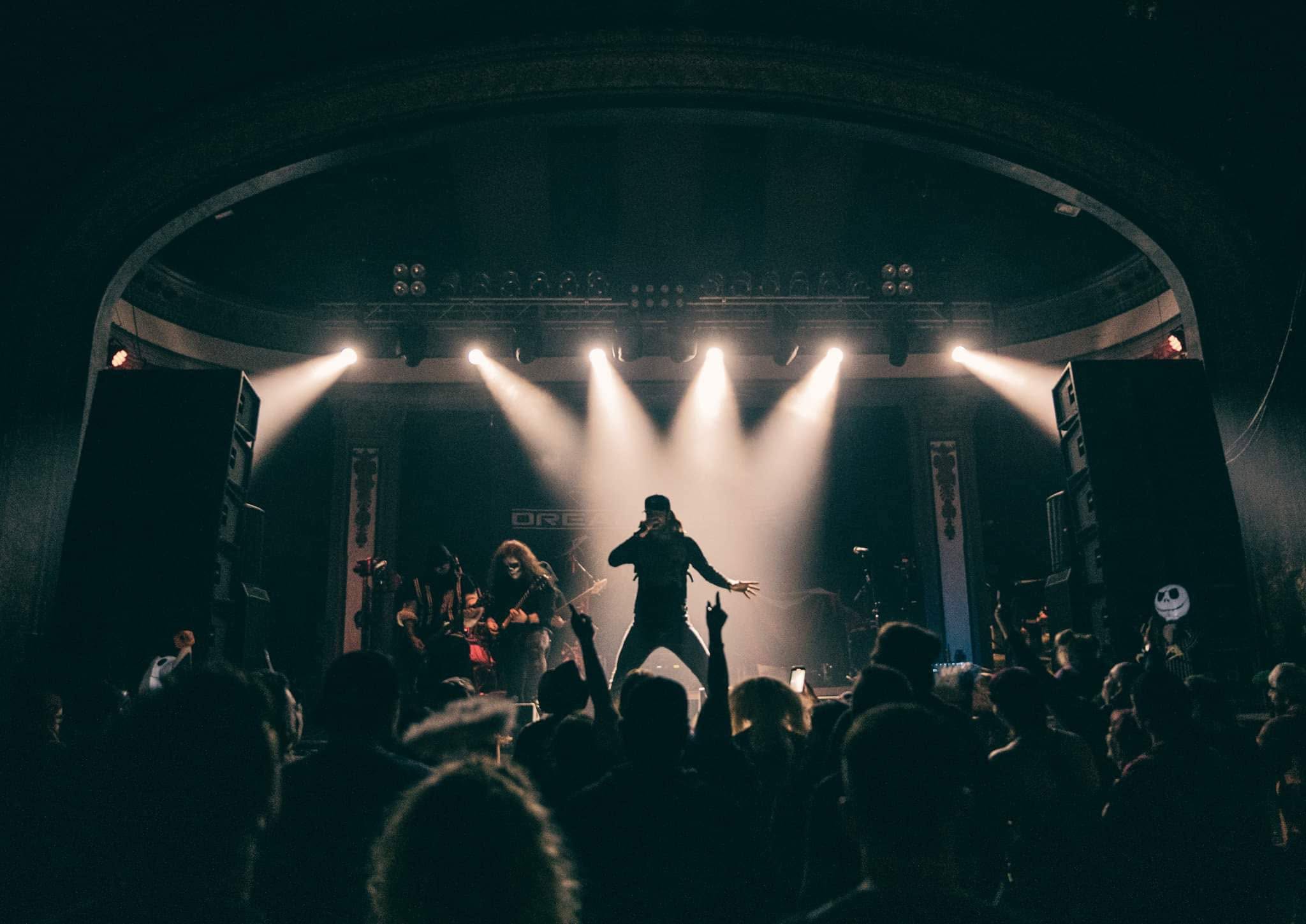 Performer on stage with bright lights, audience in foreground, concert setting, dark atmosphere.