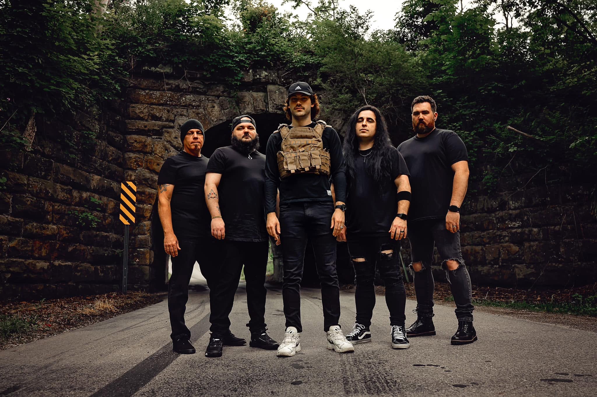 Group of five people standing on a road with a stone wall and trees behind them.