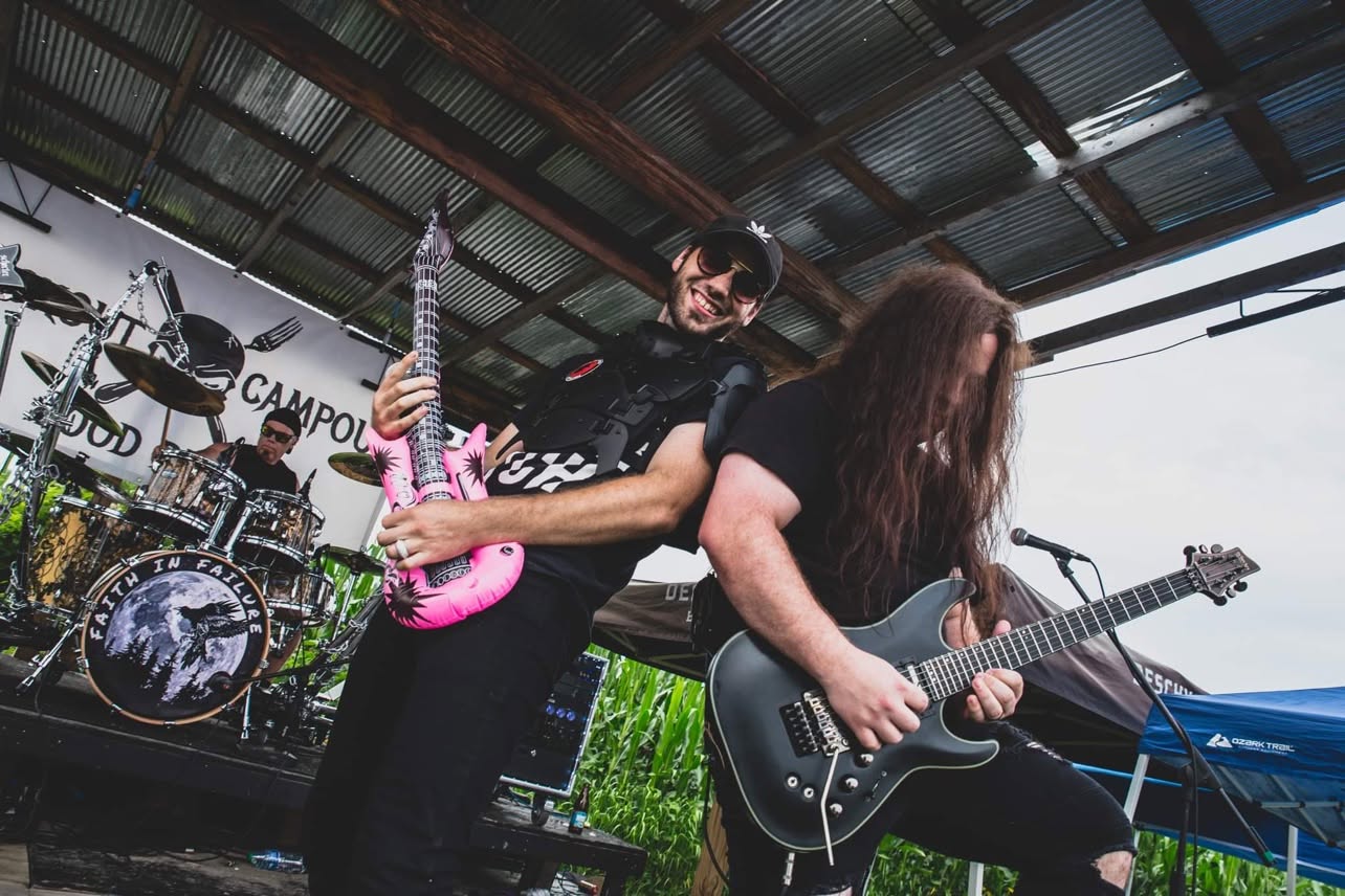 Band performing on stage with guitars and drums, under a metal roof, outdoor setting, audience visible.