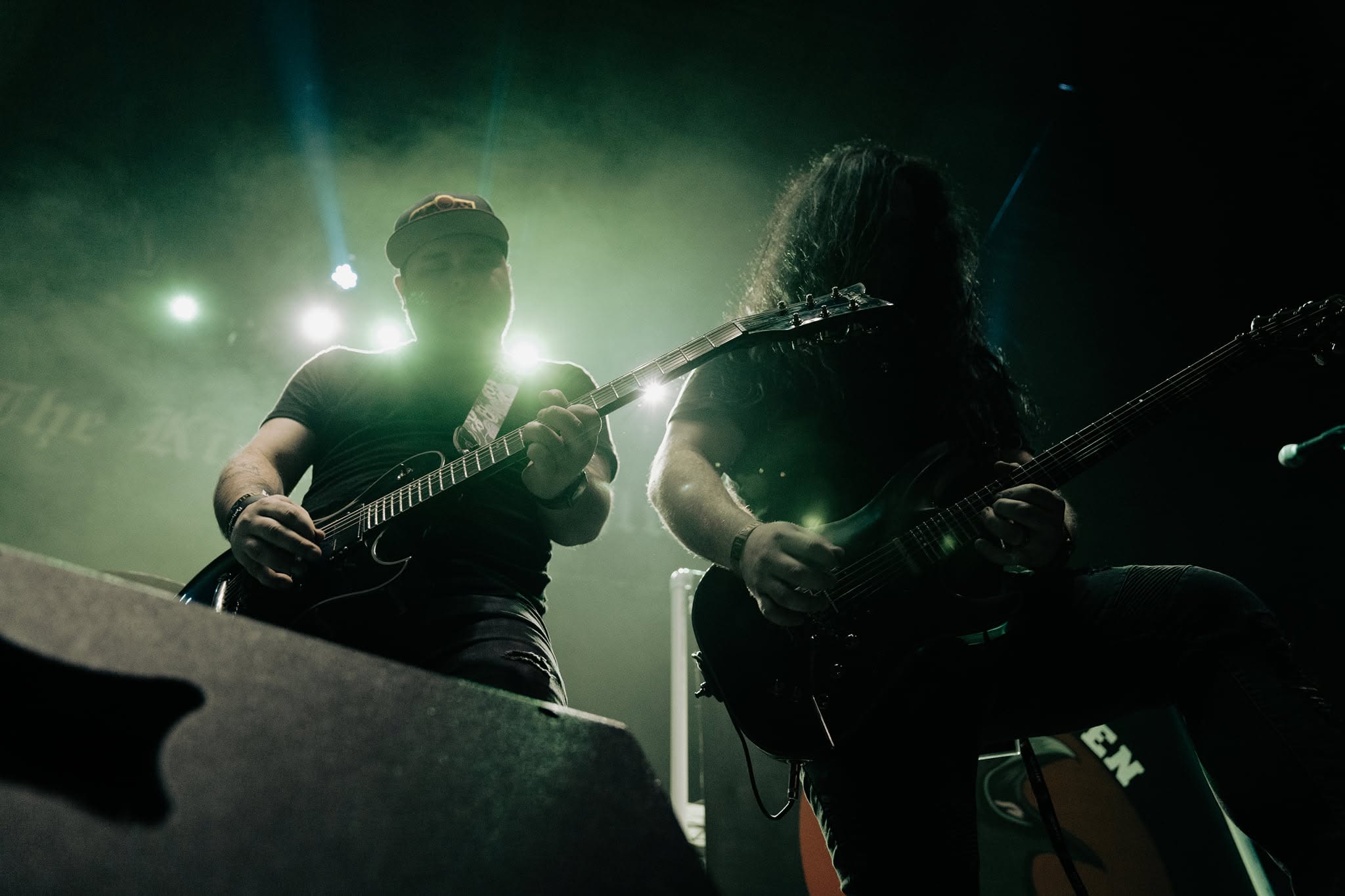 Two musicians playing electric guitars on stage with bright lights behind them, one with glasses and the other with long hair