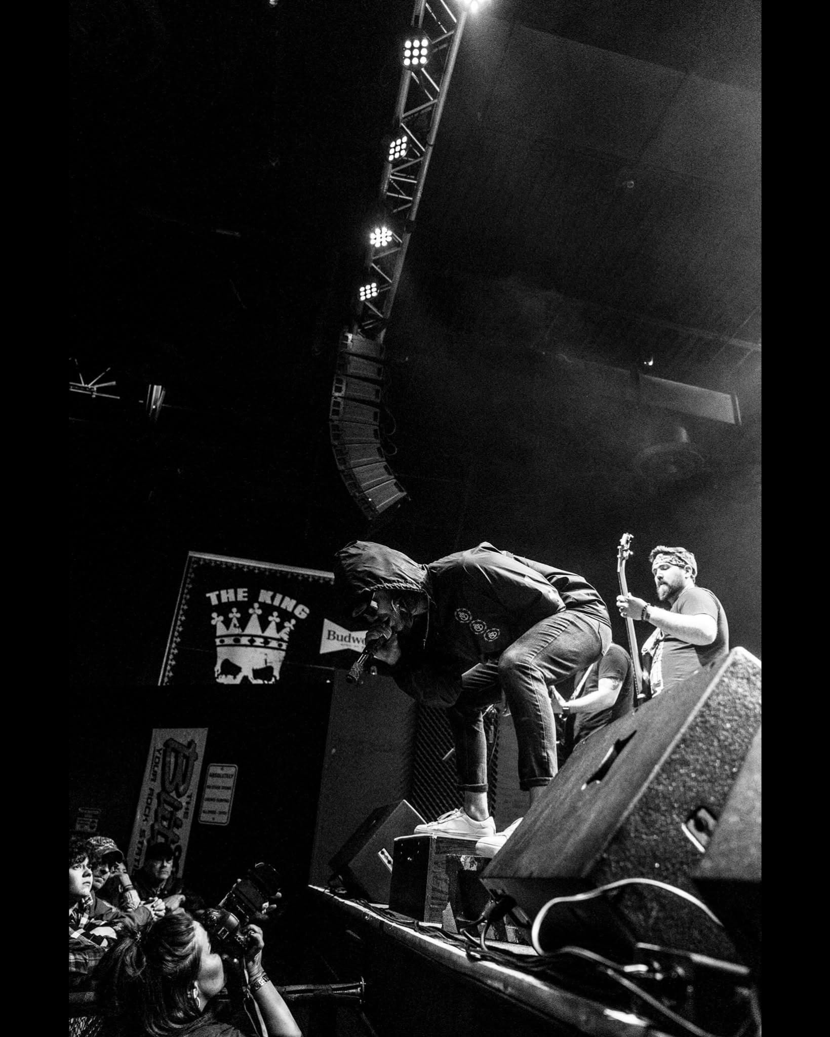People on stage adjusting equipment, audience watching, stage lights overhead, and a sign with a crown logo in the background.