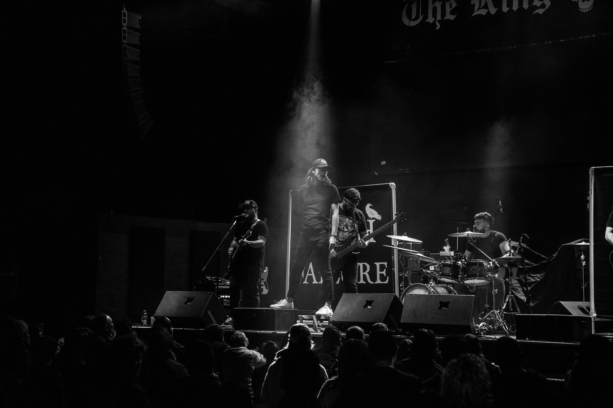 Band performs on stage with audience watching, stage lights focused on musicians, dark background, some equipment visible.