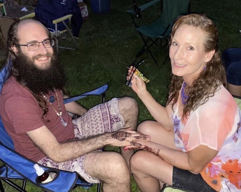 A man with glasses and a beard and a woman with curly hair sit outdoors, holding hands and smiling at each other.