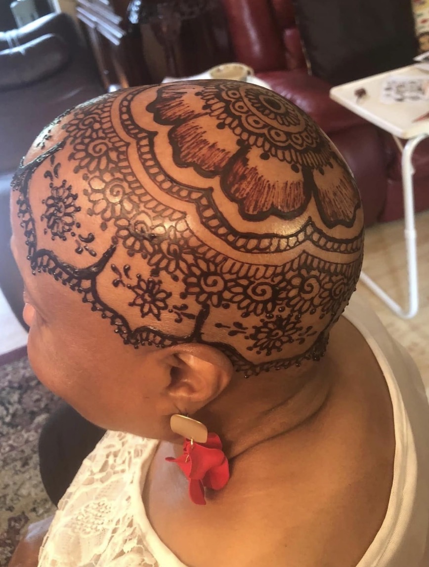 Person with a henna tattoo covering the scalp, featuring floral and geometric patterns, wearing a red earring, indoors.
