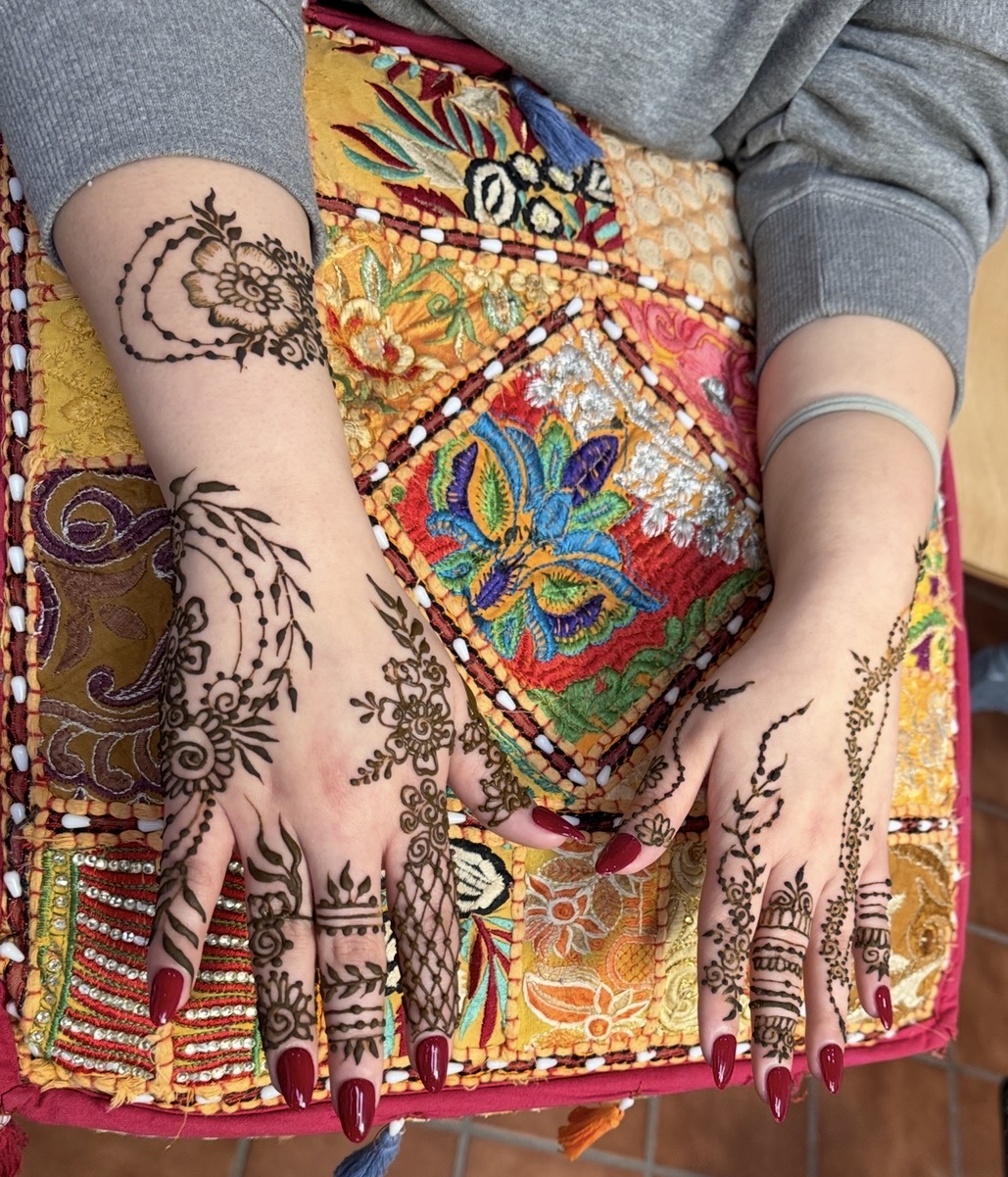 Hands with henna designs resting on colorful embroidered fabric, with long red nails and gray sleeves, no background visible.