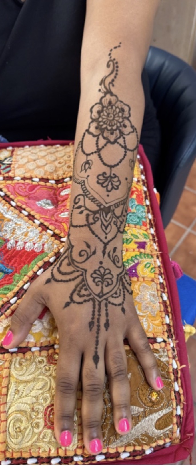 Intricate black floral and decorative tattoo on person's forearm and hand, with pink nail polish, resting on colorful fabric.