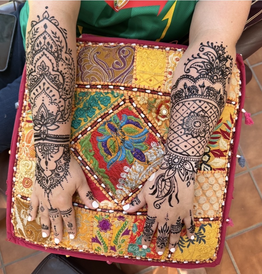 Person's hands with intricate henna designs resting on colorful embroidered fabric, arms visible, background partially visible.