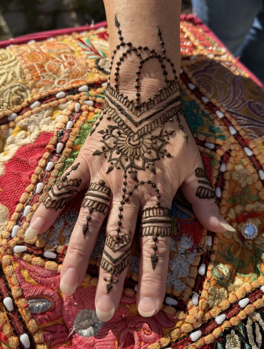 Hand with intricate henna designs resting on colorful fabric, detailed patterns visible on fingers and wrist.