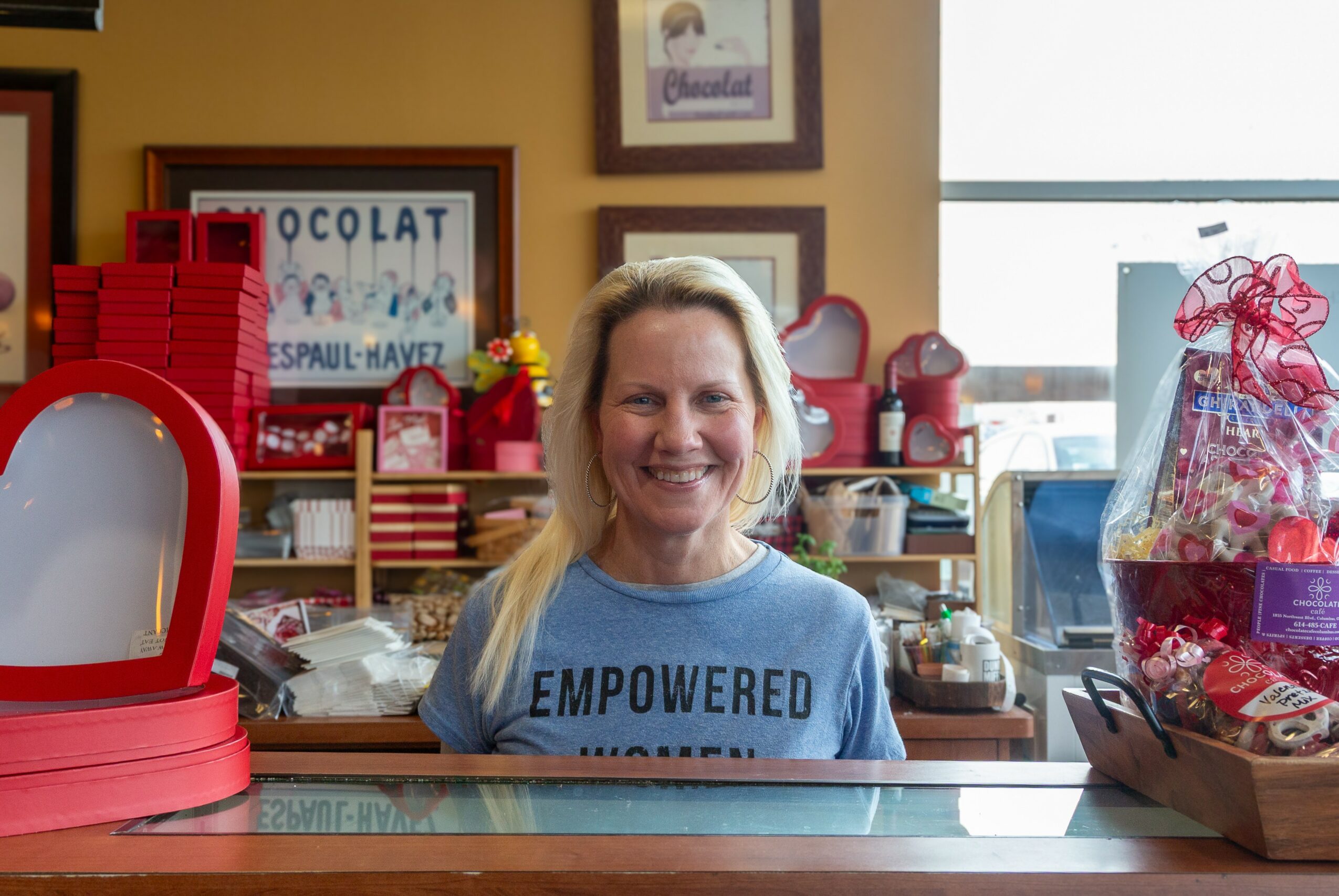 Woman smiling behind a counter with gift baskets and colorful items, window in background