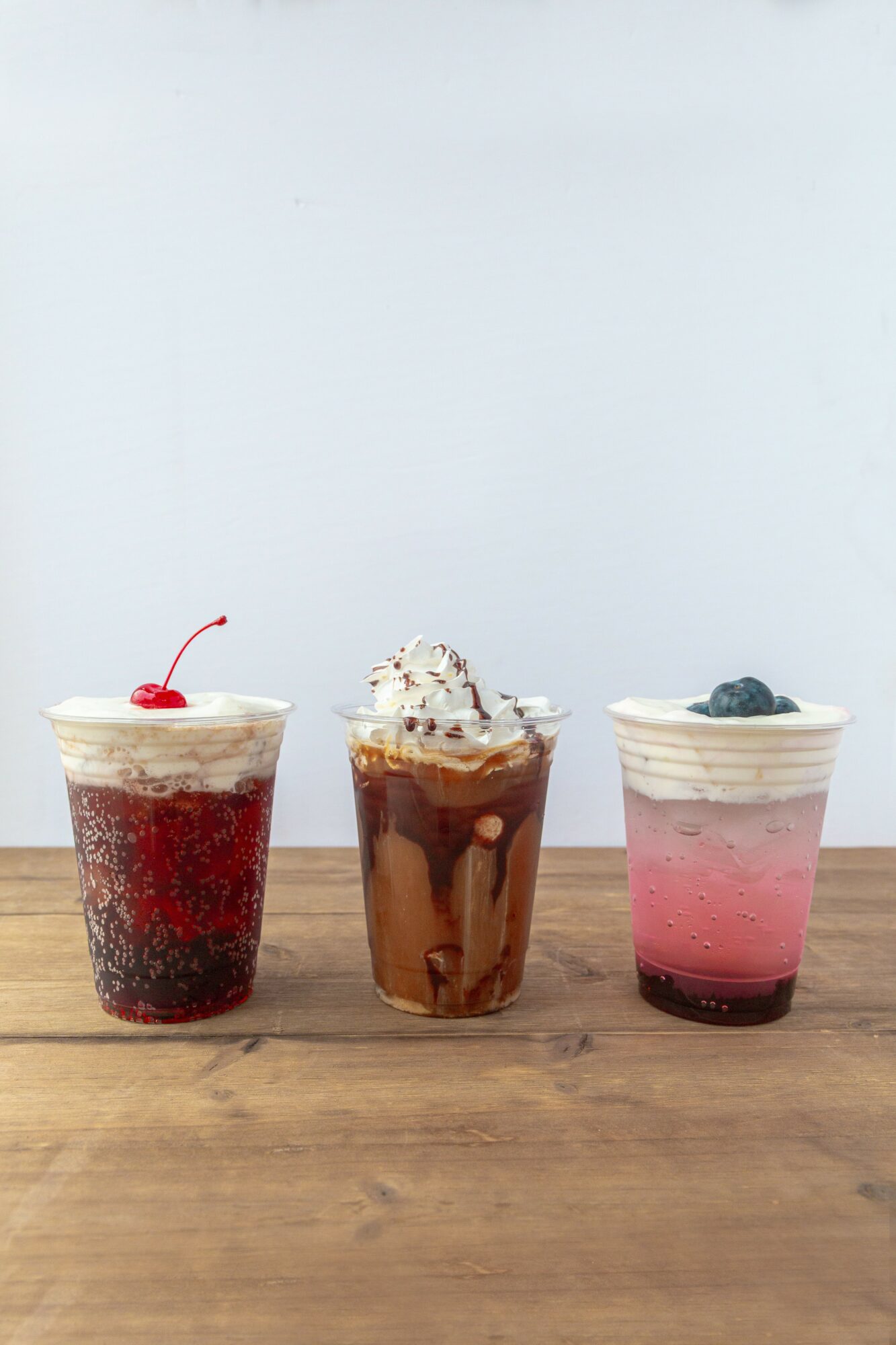 Three colorful beverages in cups with toppings on a wooden surface against a plain background.