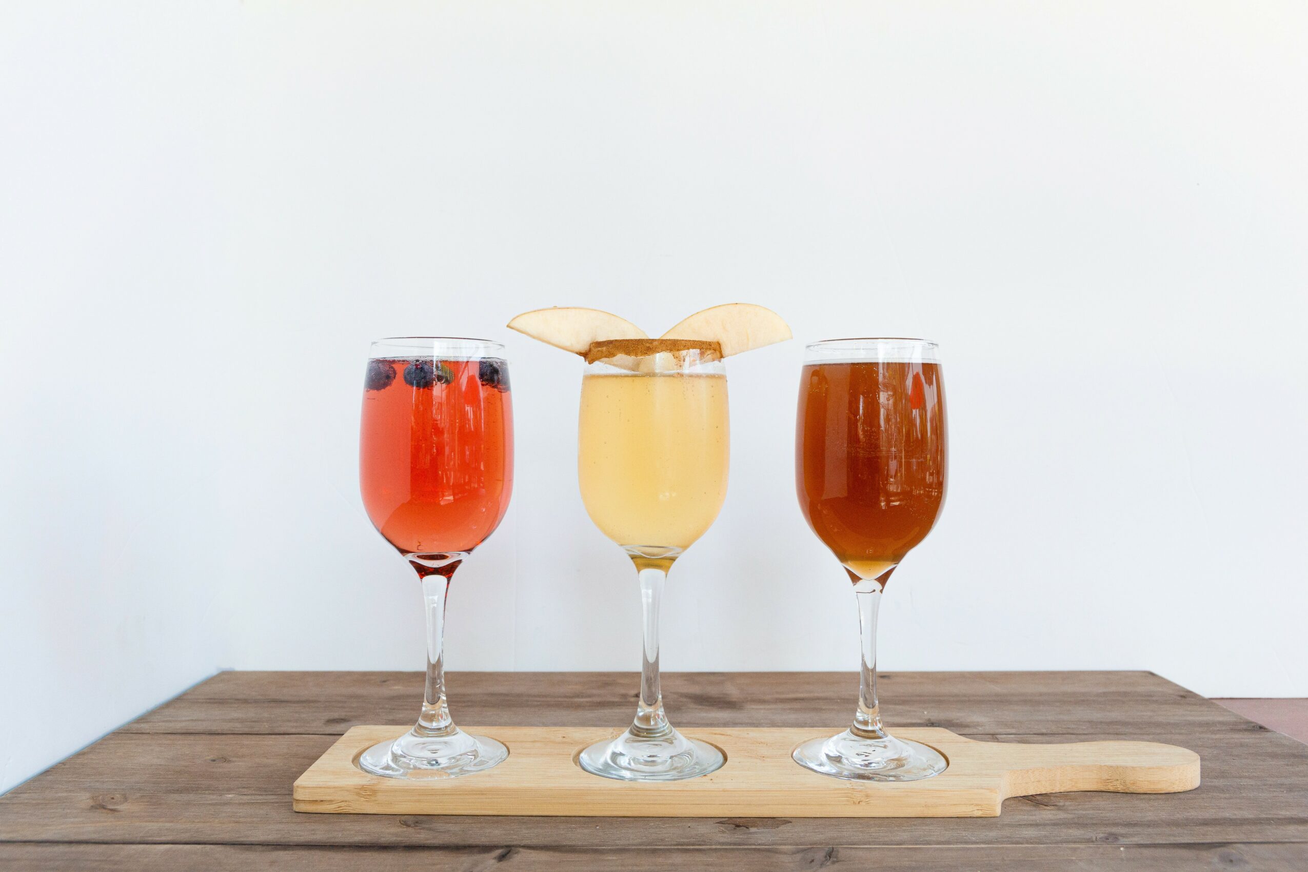 Three colorful cocktails on a wooden board, with a plain light background, each in a tall glass with different garnishes.