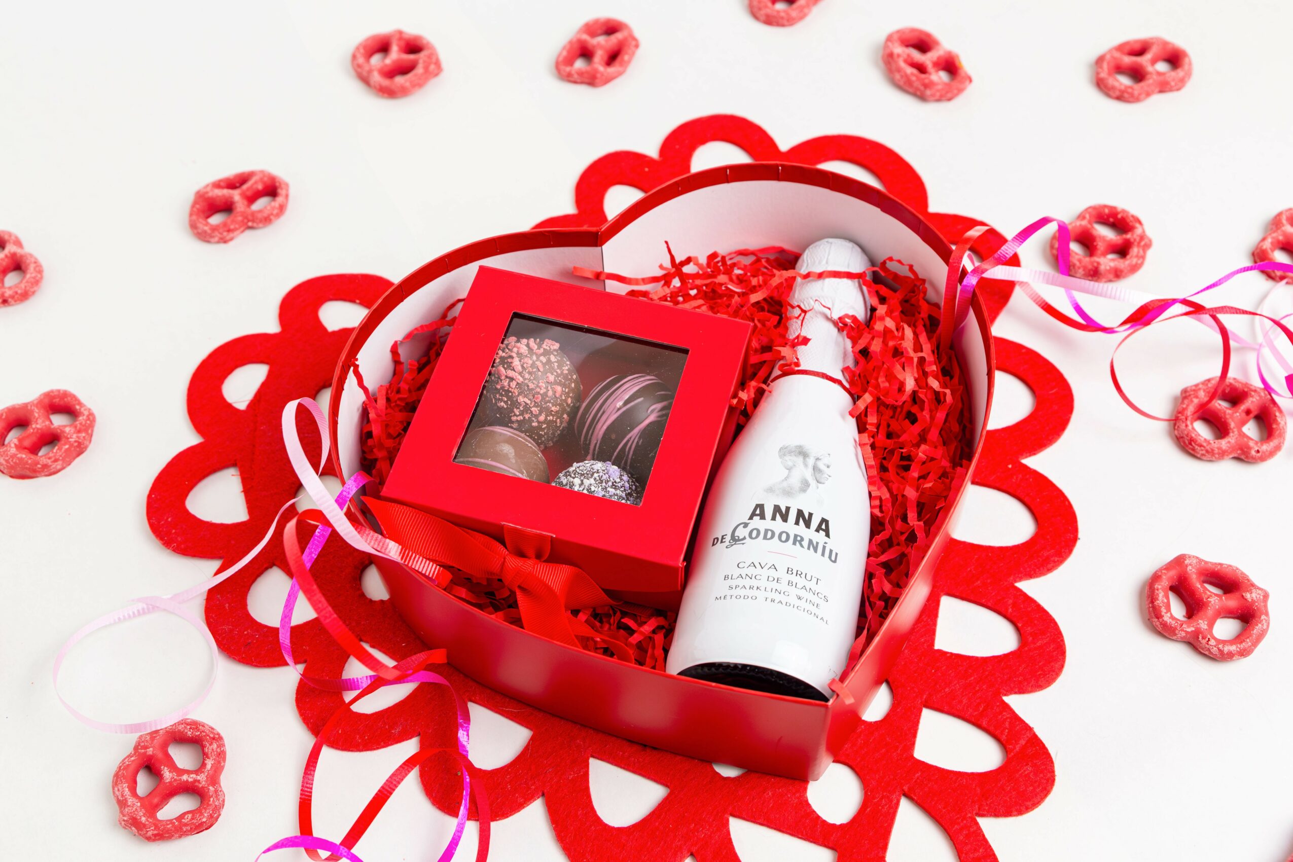 Red heart-shaped box with chocolates, a white bottle, and pink ribbons, surrounded by small pretzel-shaped candies on a white background.