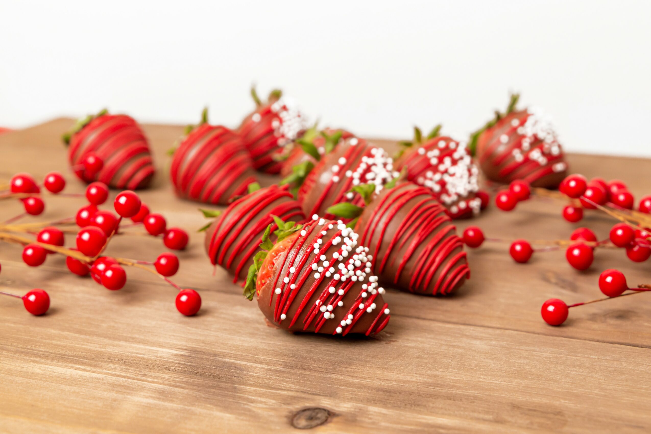 Strawberries dipped in chocolate with white sprinkles, surrounded by red berries on a wooden surface.