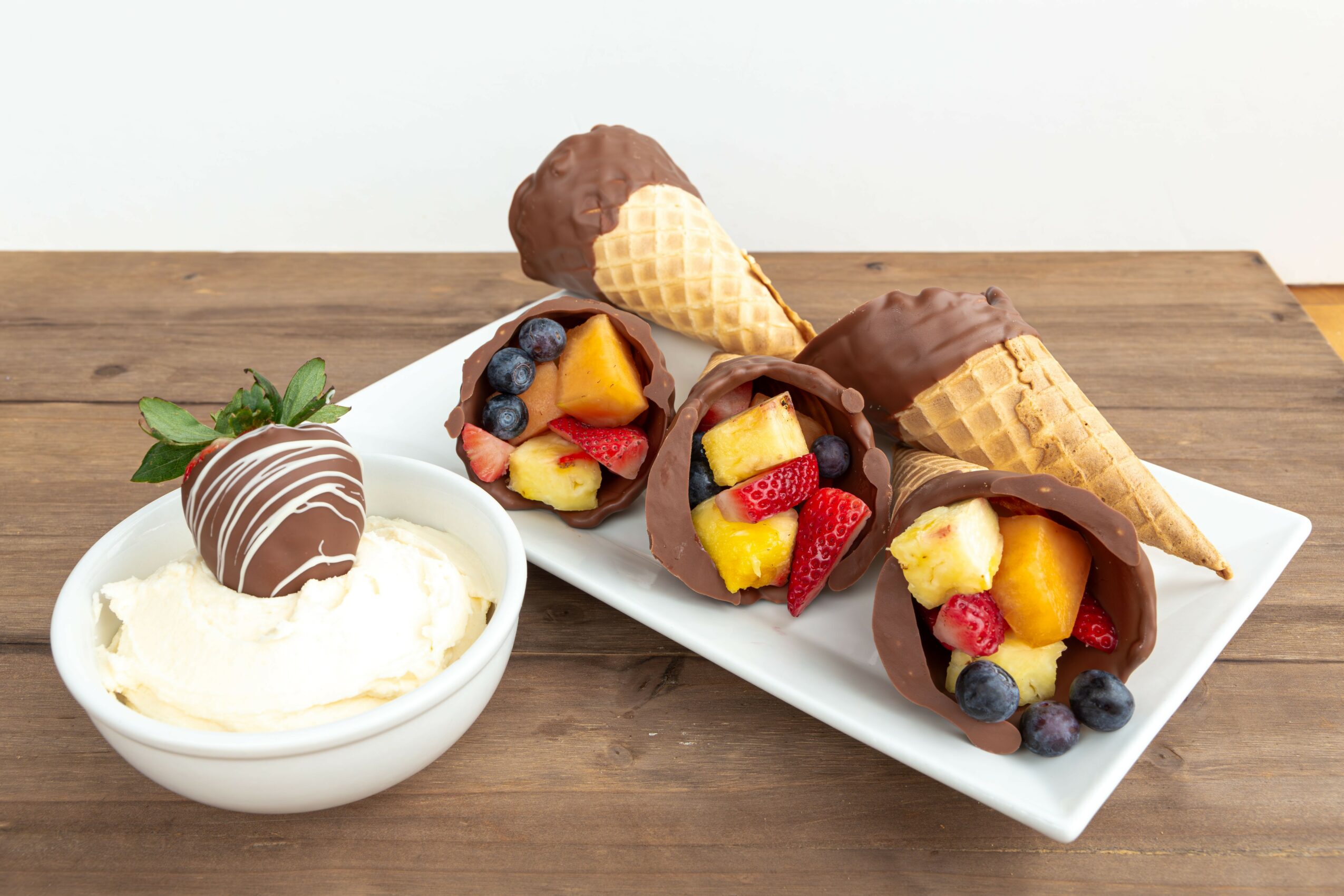 Three chocolate-dipped ice cream cones with fruit filling on a white plate, beside a bowl of vanilla ice cream with a chocolate-covered scoop, on a wooden surface.