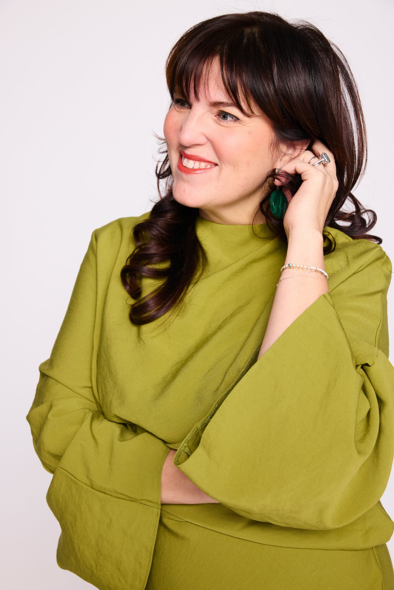 Woman with dark hair smiling, wearing a green top, touching her ear, against a plain white background.