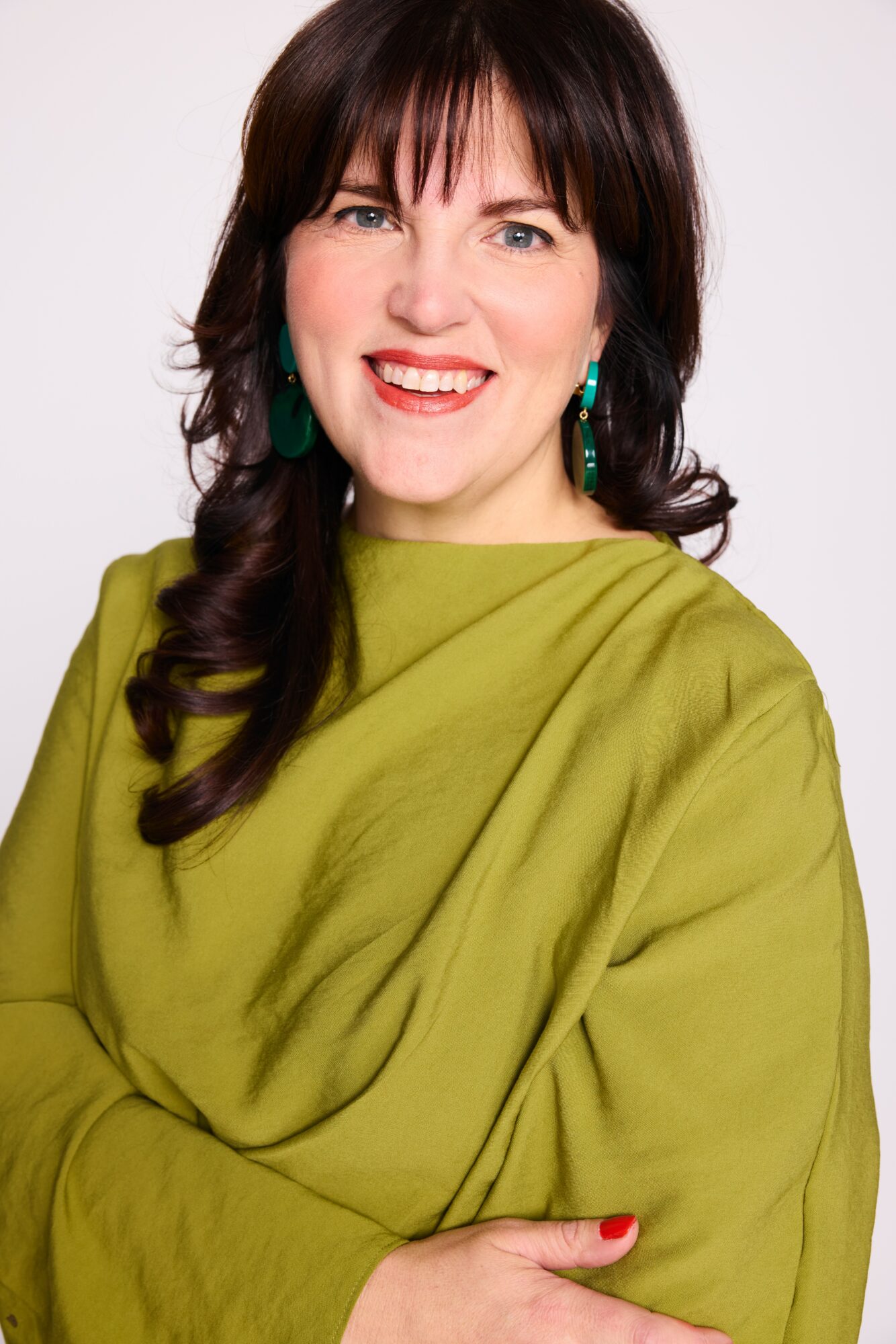 Woman with dark hair, smiling, wearing green earrings and a green top, against a white background.