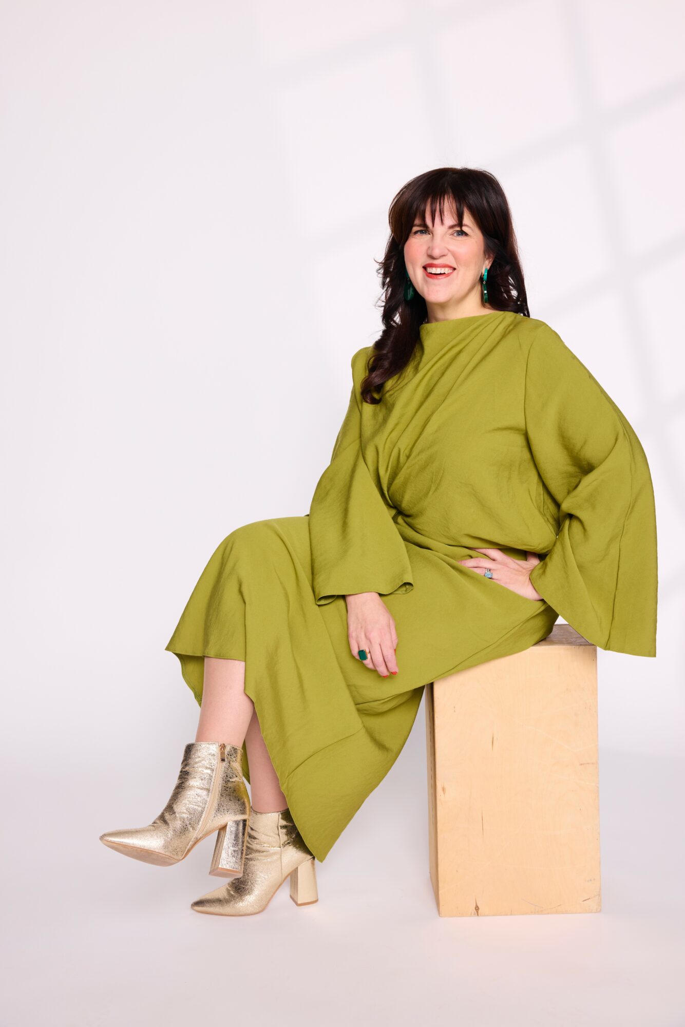 Woman in a green dress sitting on a wooden block, smiling, with a white background and shadow pattern.