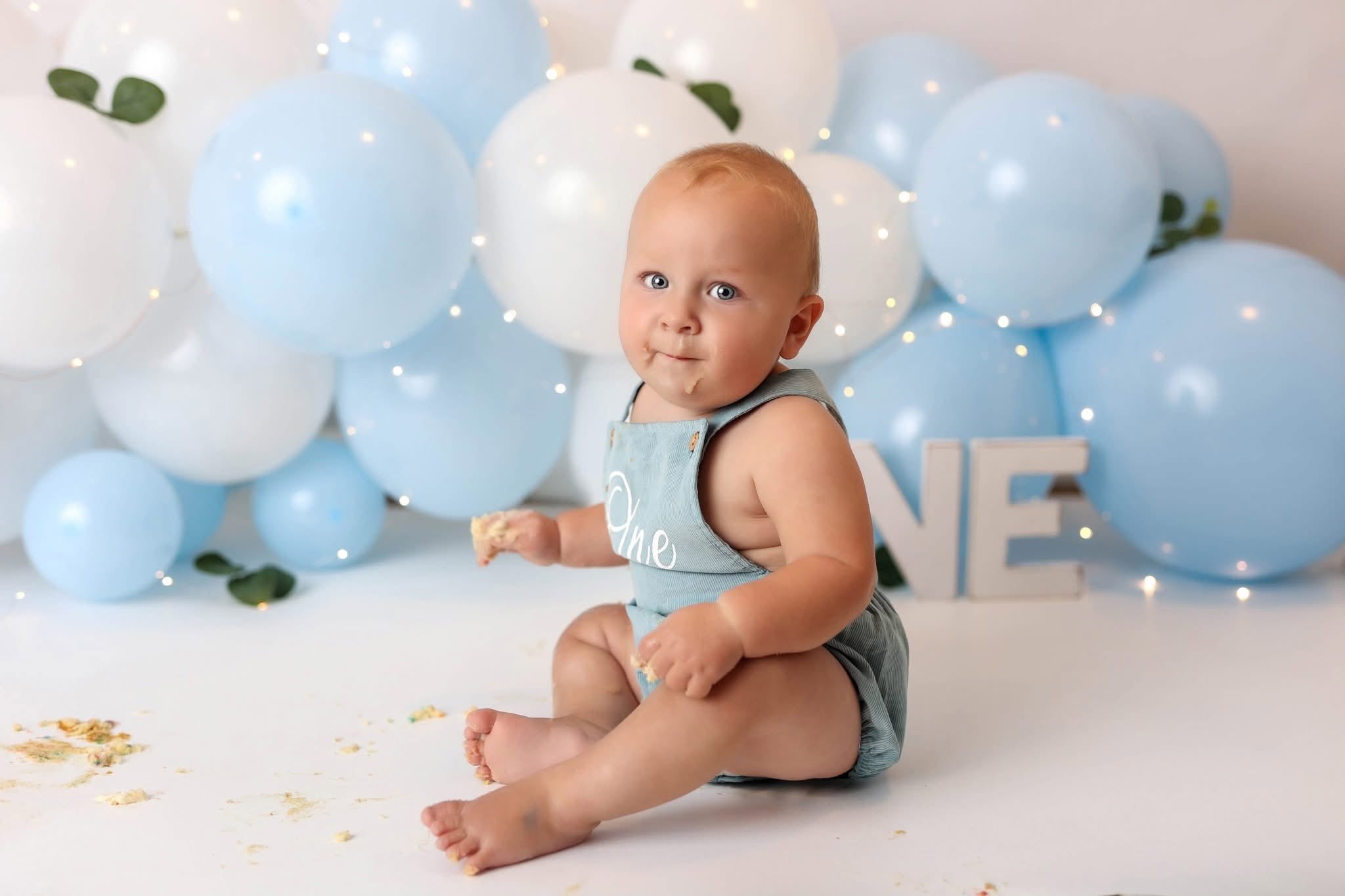 Baby sitting on floor with blue and white balloons behind, wearing a light blue outfit, looking at camera.