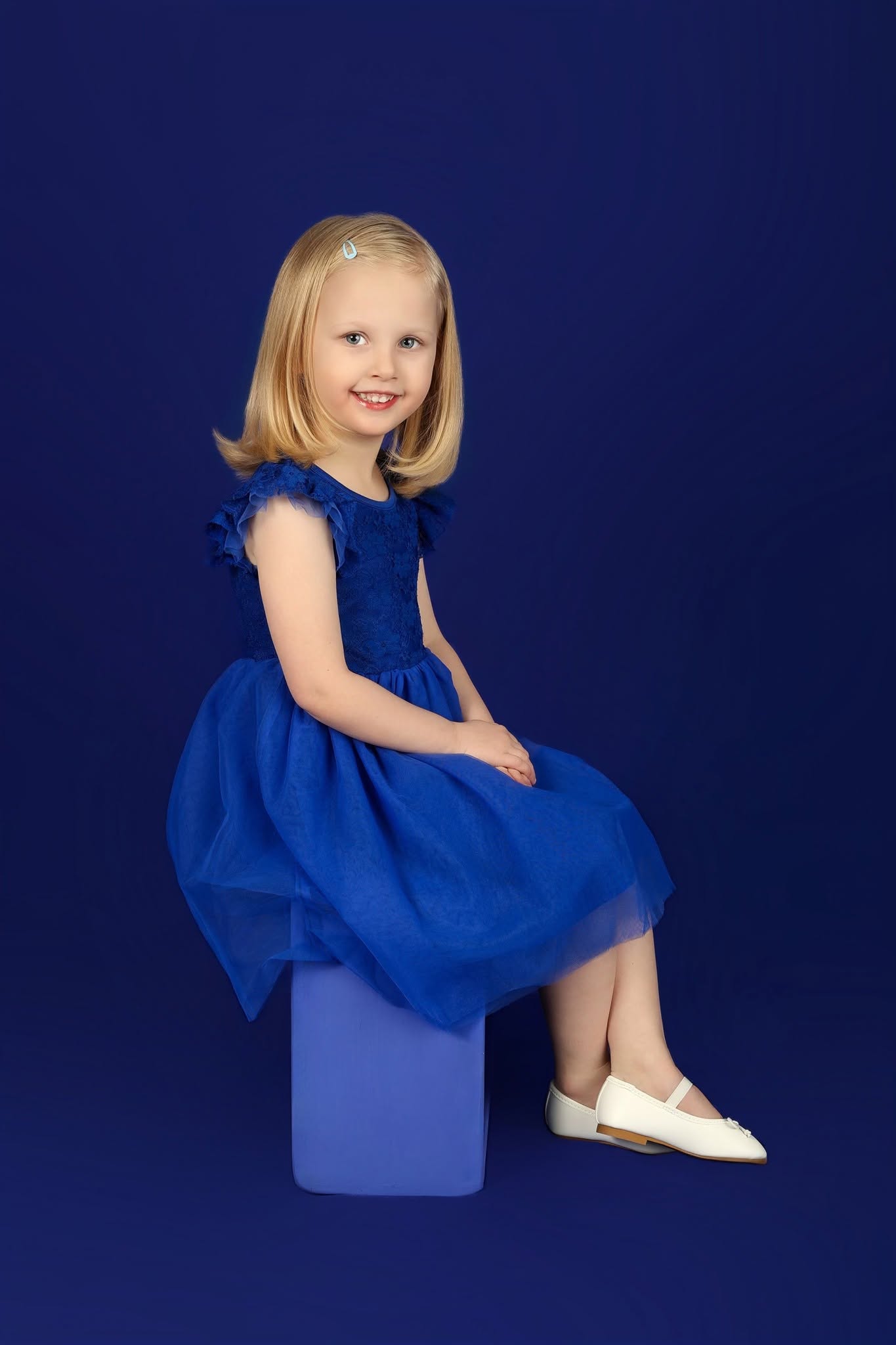 Young girl with blonde hair in a blue dress sitting on a blue block against a dark blue background, smiling.