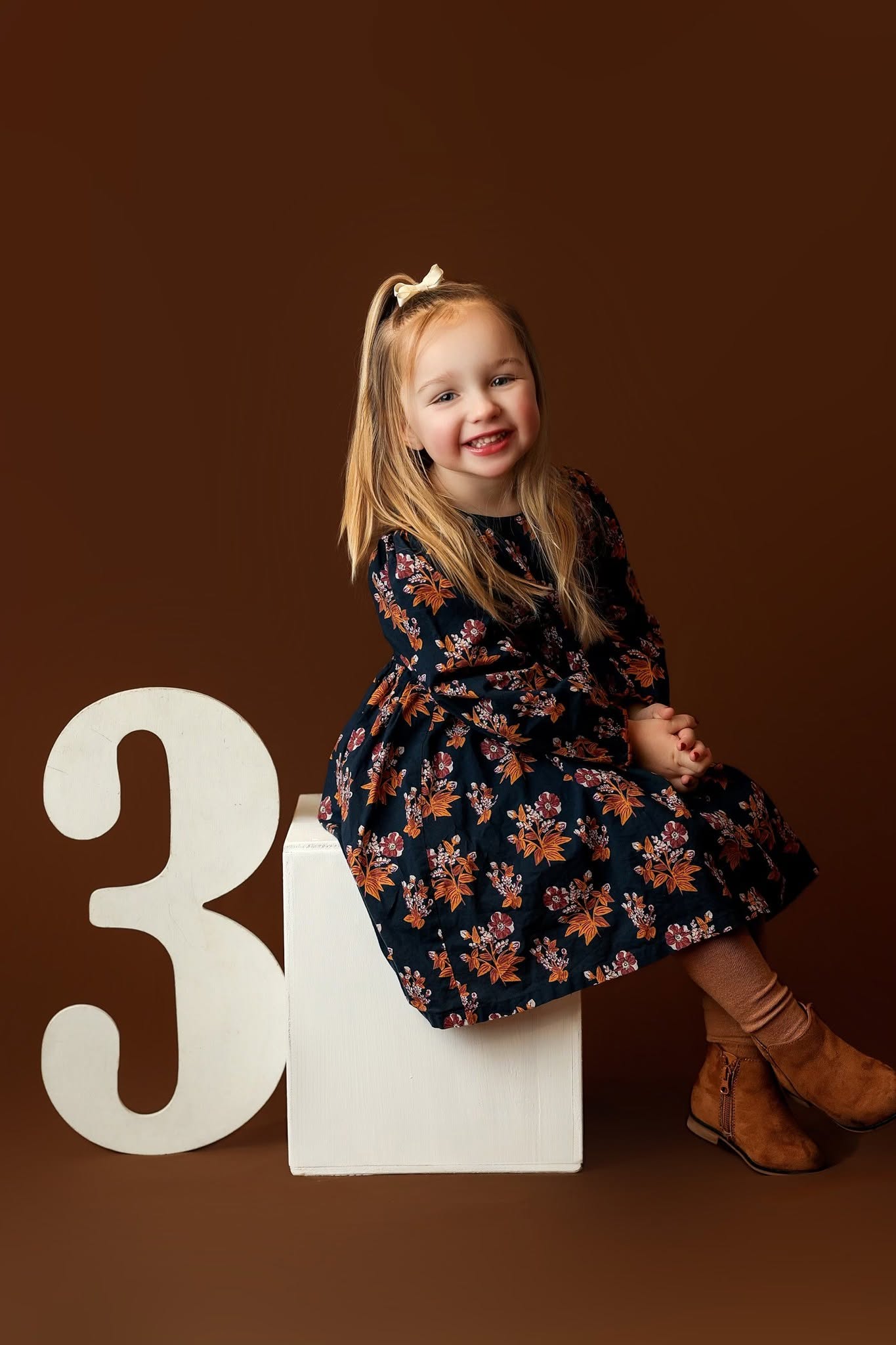 Young girl with long blonde hair, wearing a floral dress and brown boots, sitting on a white block, smiling.