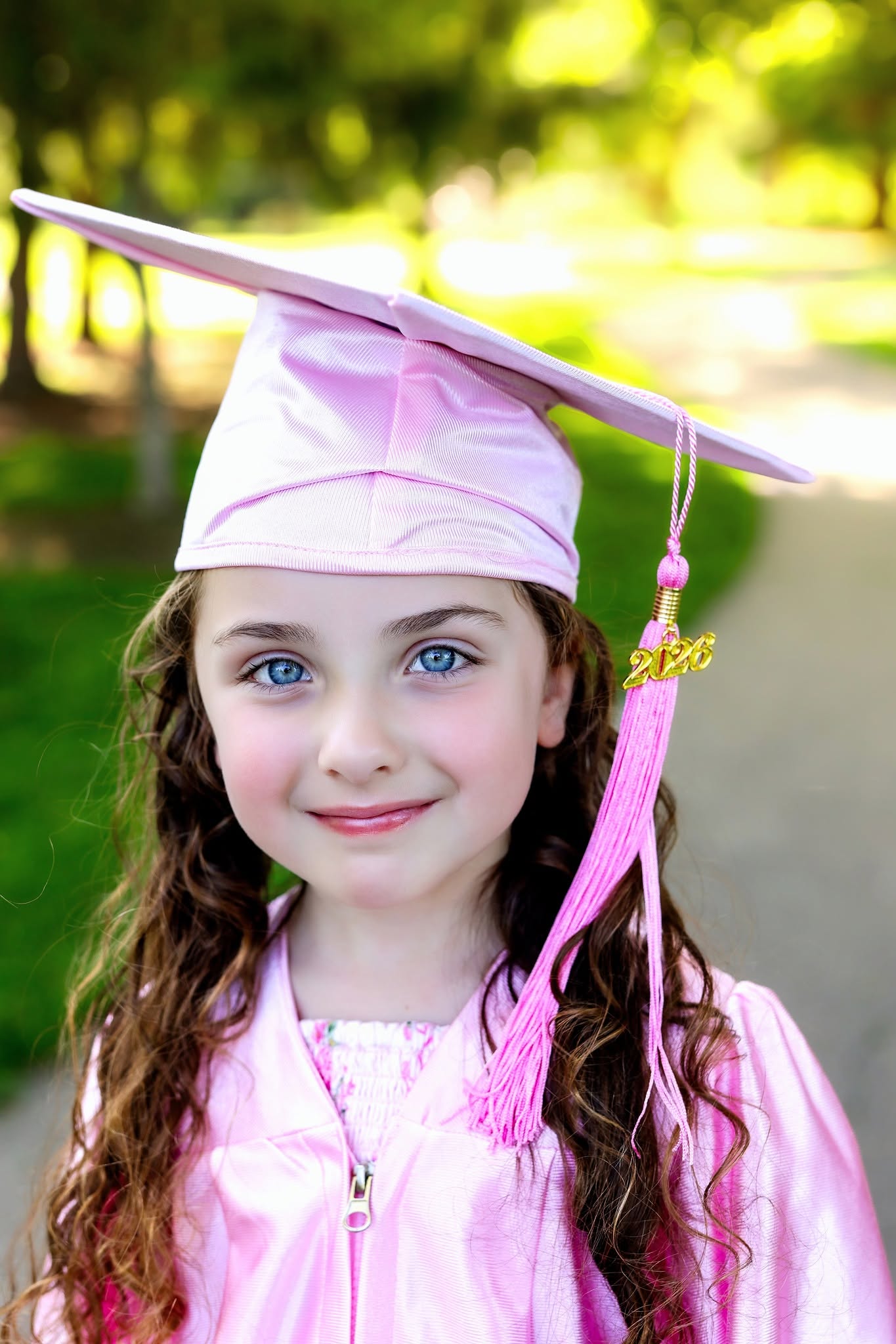 Young girl in pink graduation cap and gown outdoors, smiling with trees in background.