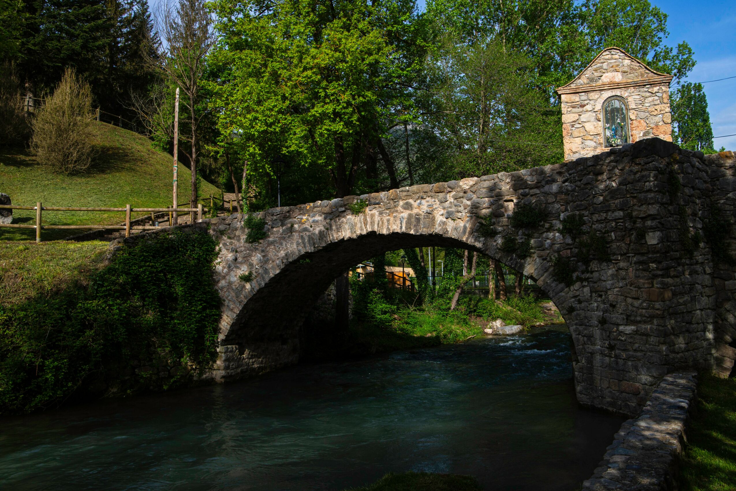 Stone bridge over a river with trees and a small building in the background.