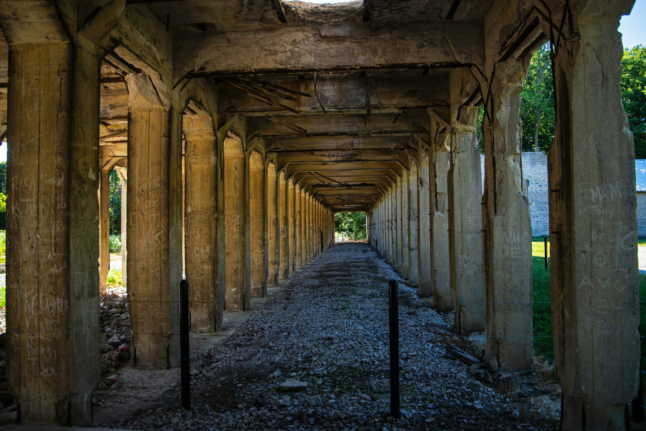 Underground passageway with wooden beams and columns, gravel path, and sunlight at the end.