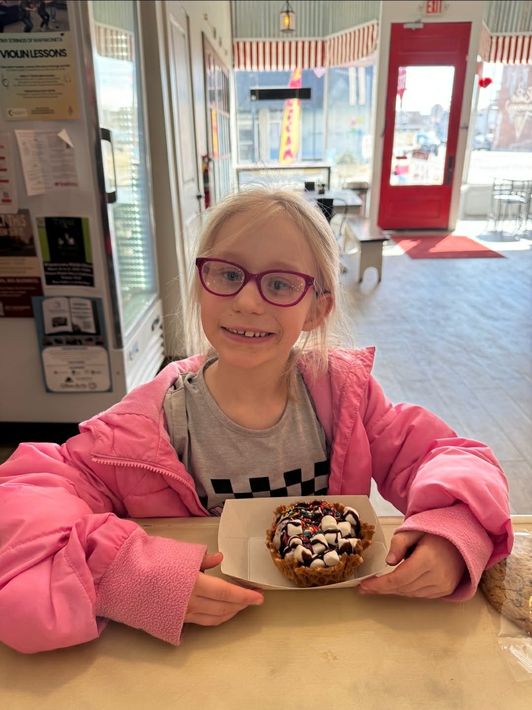 Young girl with glasses and a pink jacket holds a decorated cupcake inside a cafe or shop.
