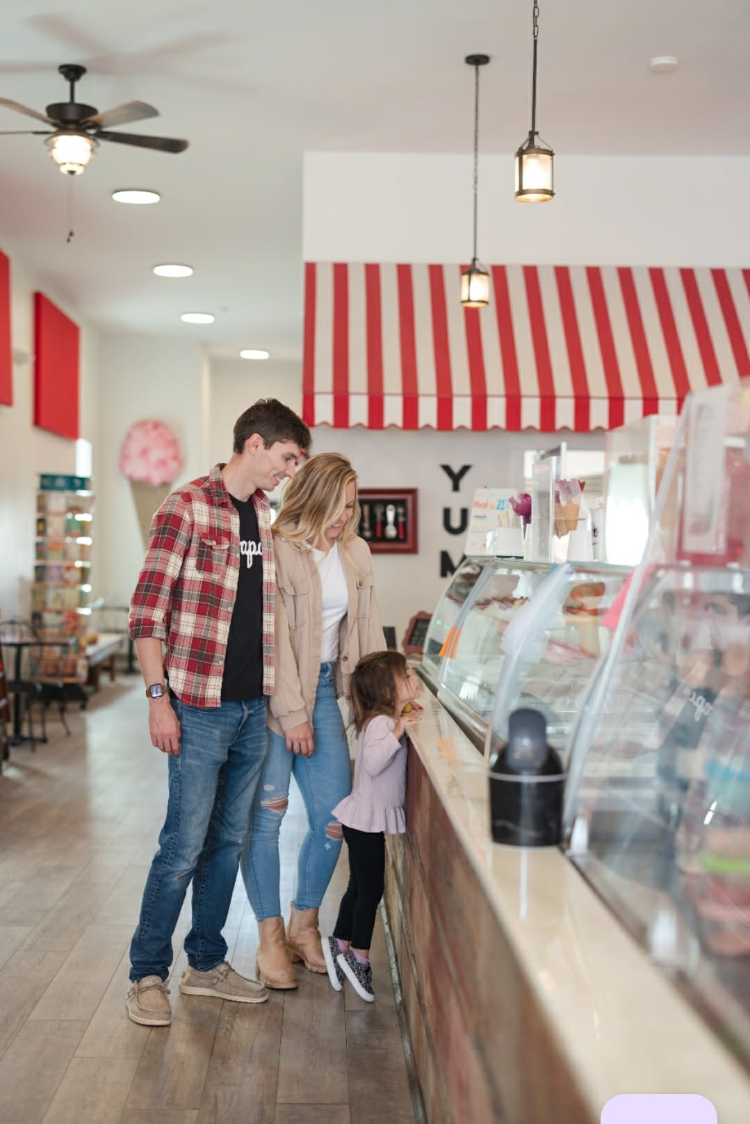 Group of three people and a child at an ice cream shop counter, with red and white striped awning, ceiling fan, and hanging lights.