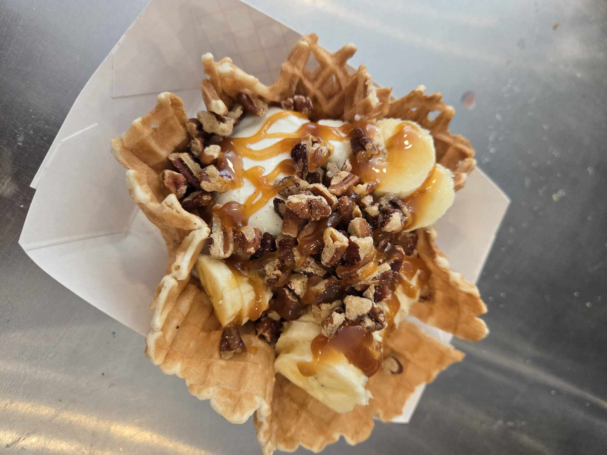 Waffle cup filled with bananas, chopped nuts, caramel drizzle, and chocolate syrup, on a white paper liner.