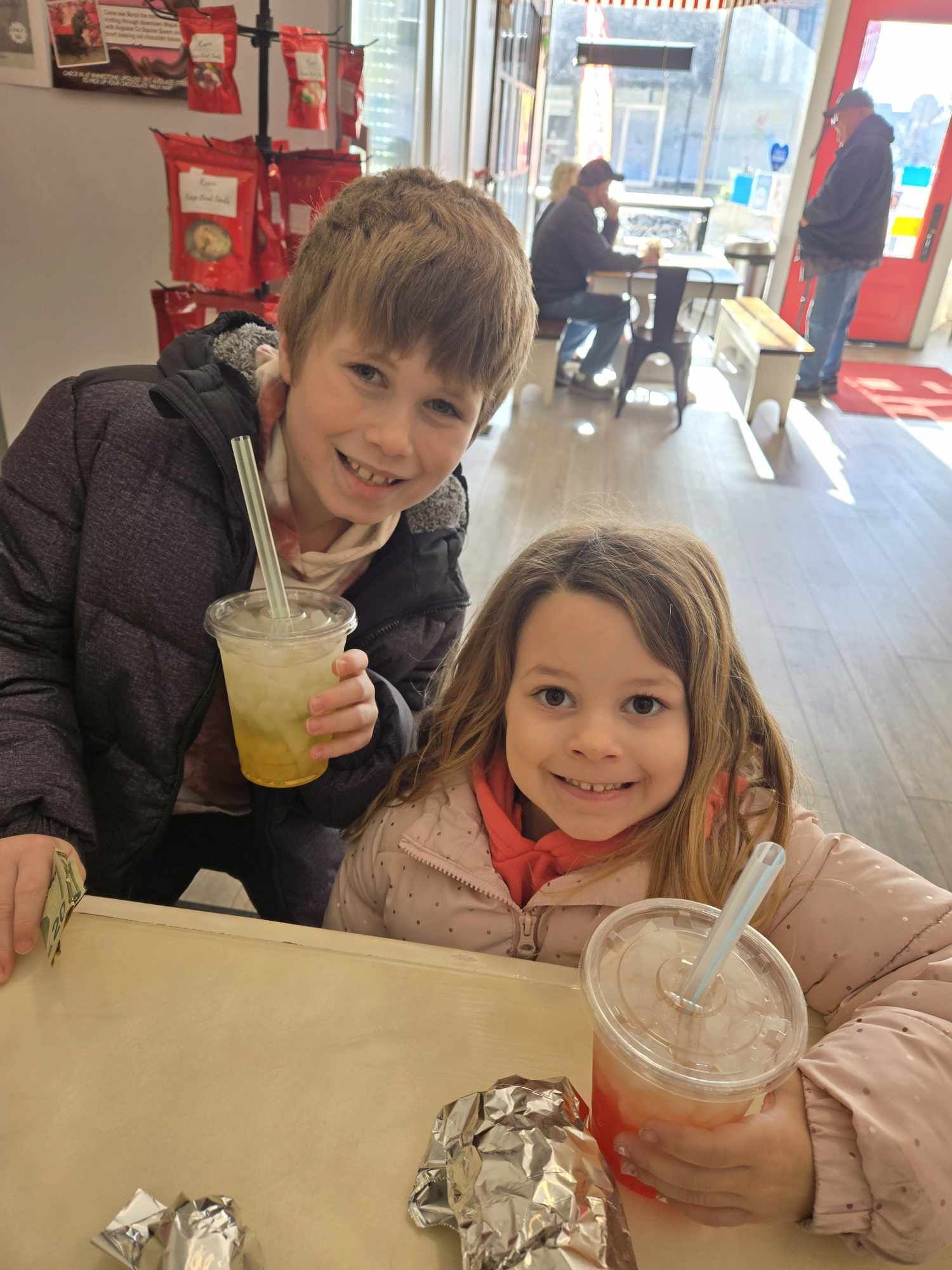 Two children smiling at a table inside a store, holding drinks with straws, with other customers in the background.