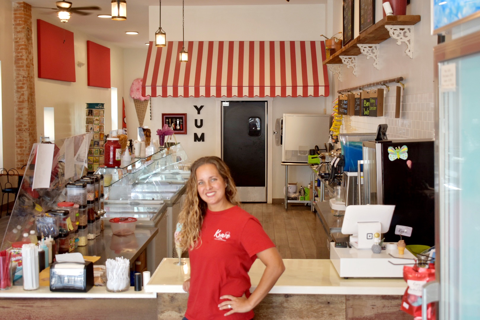 Smiling woman in red shirt behind a counter in a cafe or bakery, with shelves and equipment visible.