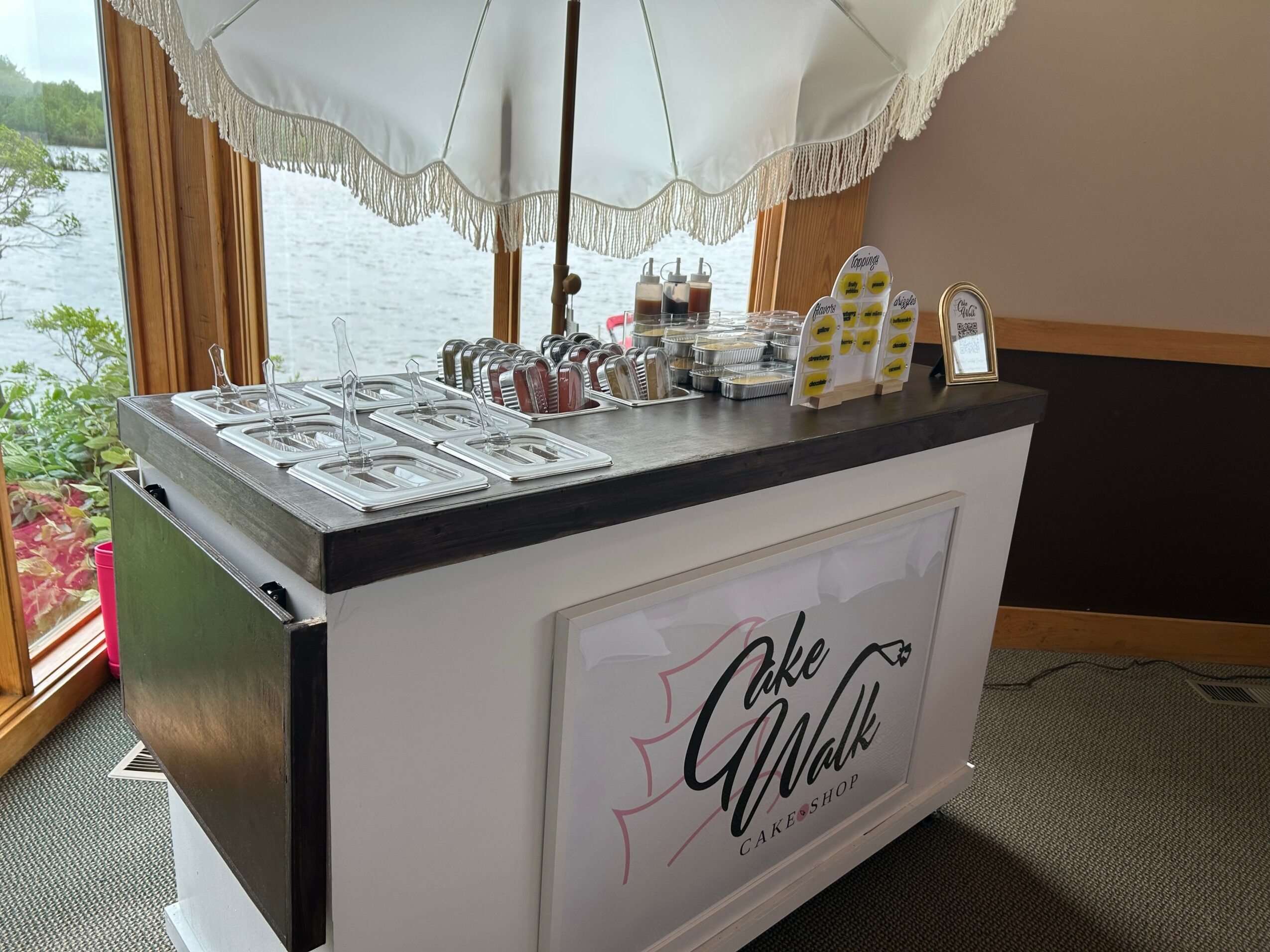 Ice cream or dessert station with toppings under a white umbrella, with a sign reading 'Cake Walk' on the front.